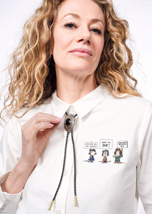 S=Close up of person with wavy blonde hair holding collar of The Peanuts x Wildfang Long Sleeve Button Up, which is a white long sleeve button up shirt with Peanuts characters embroidery on right chest.