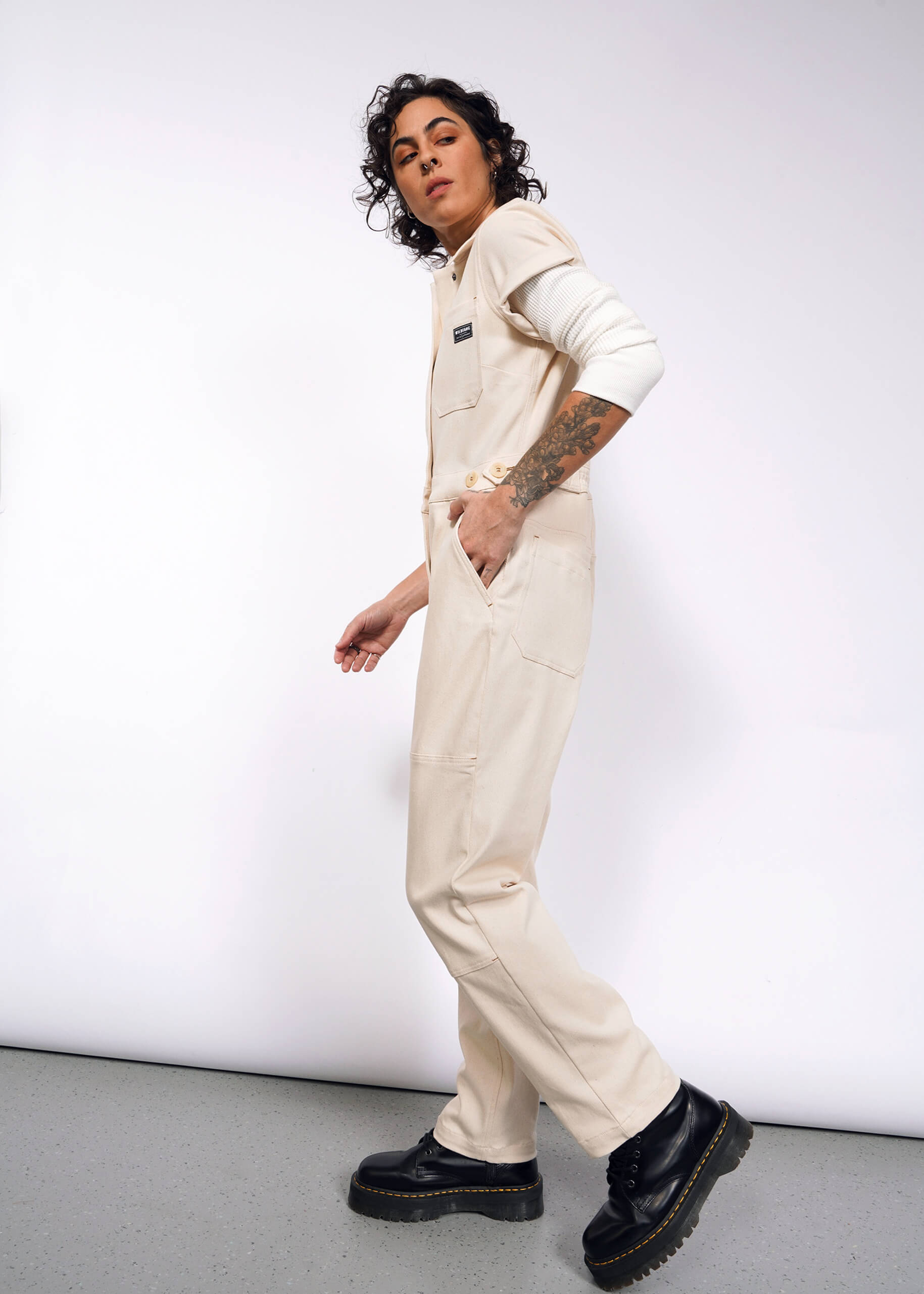 S=A person with curly hair models The Essential Denim High Waisted Coverall, featuring deep pockets and an undyed finish. Tattoos adorn their left arm as they tuck their right hand casually. They wear black shoes while appearing in motion against a plain backdrop.