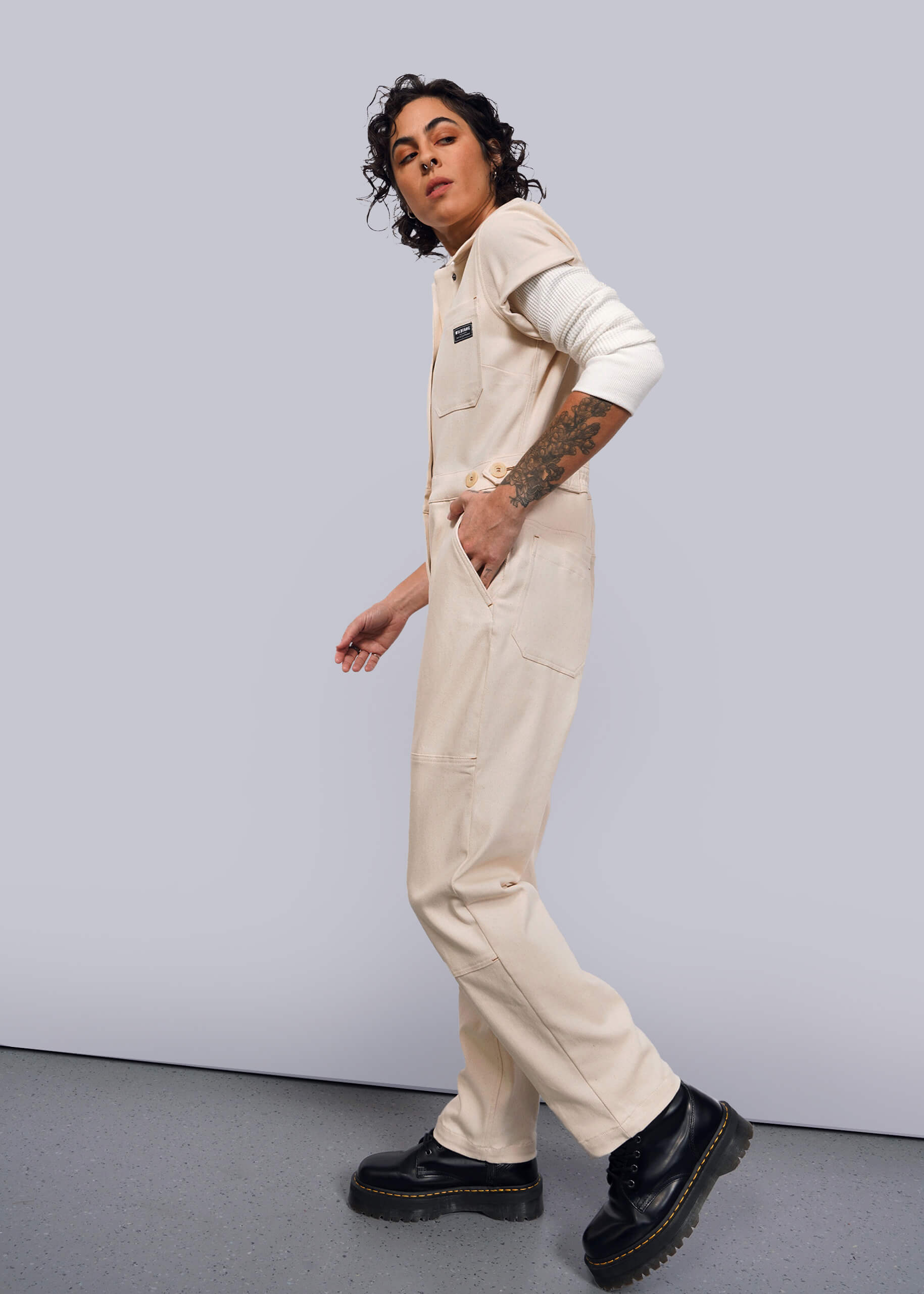 S=A person with curly hair models The Essential Denim High Waisted Coverall, featuring deep pockets and an undyed finish. Tattoos adorn their left arm as they tuck their right hand casually. They wear black shoes while appearing in motion against a plain backdrop.