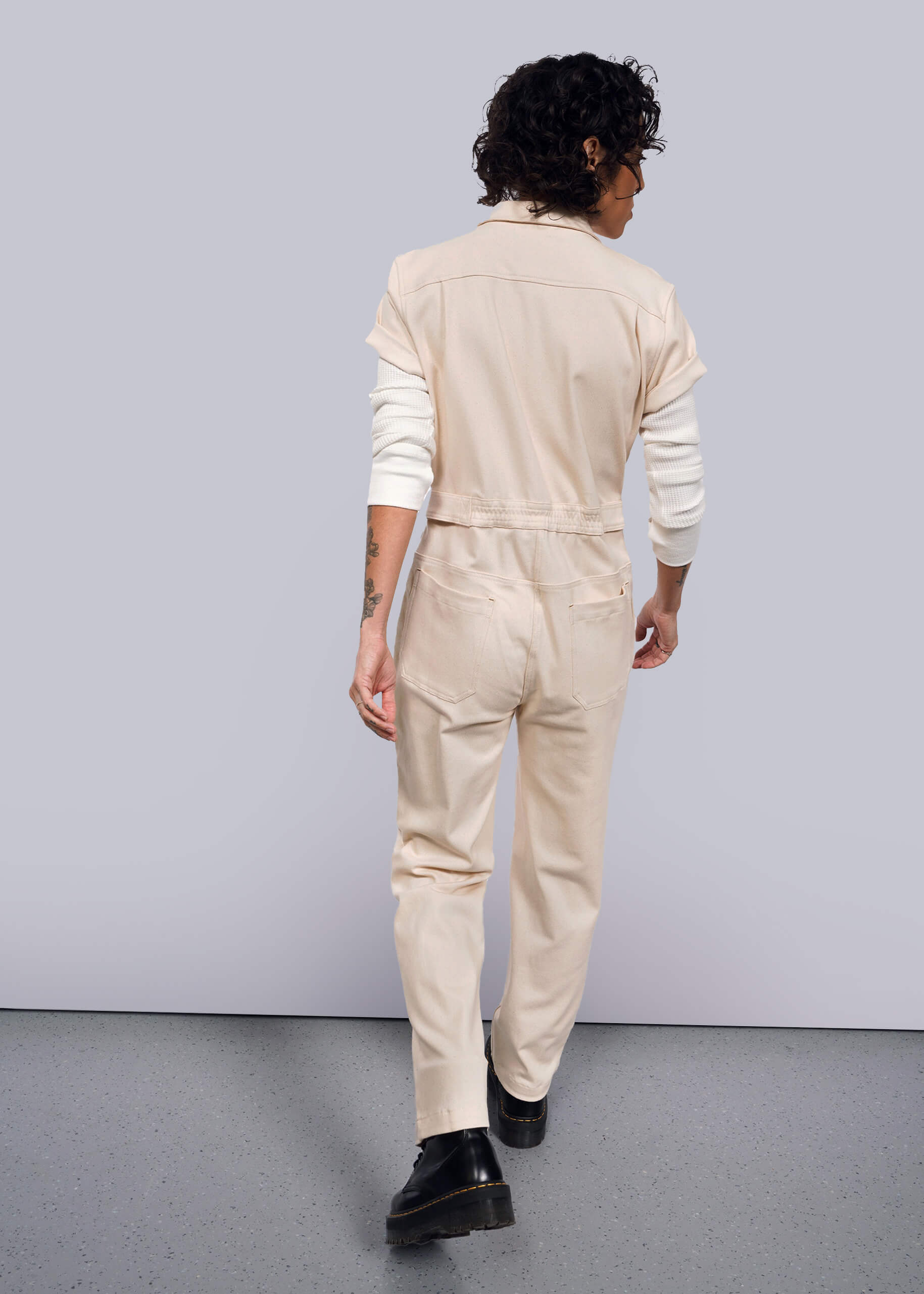 S= Facing away, a person with curly hair wears The Essential Denim High Waisted Coverall in cream over a long-sleeve white shirt, displaying tattoos on their arm. They walk on a speckled floor against a white backdrop, completing the look with black shoes.