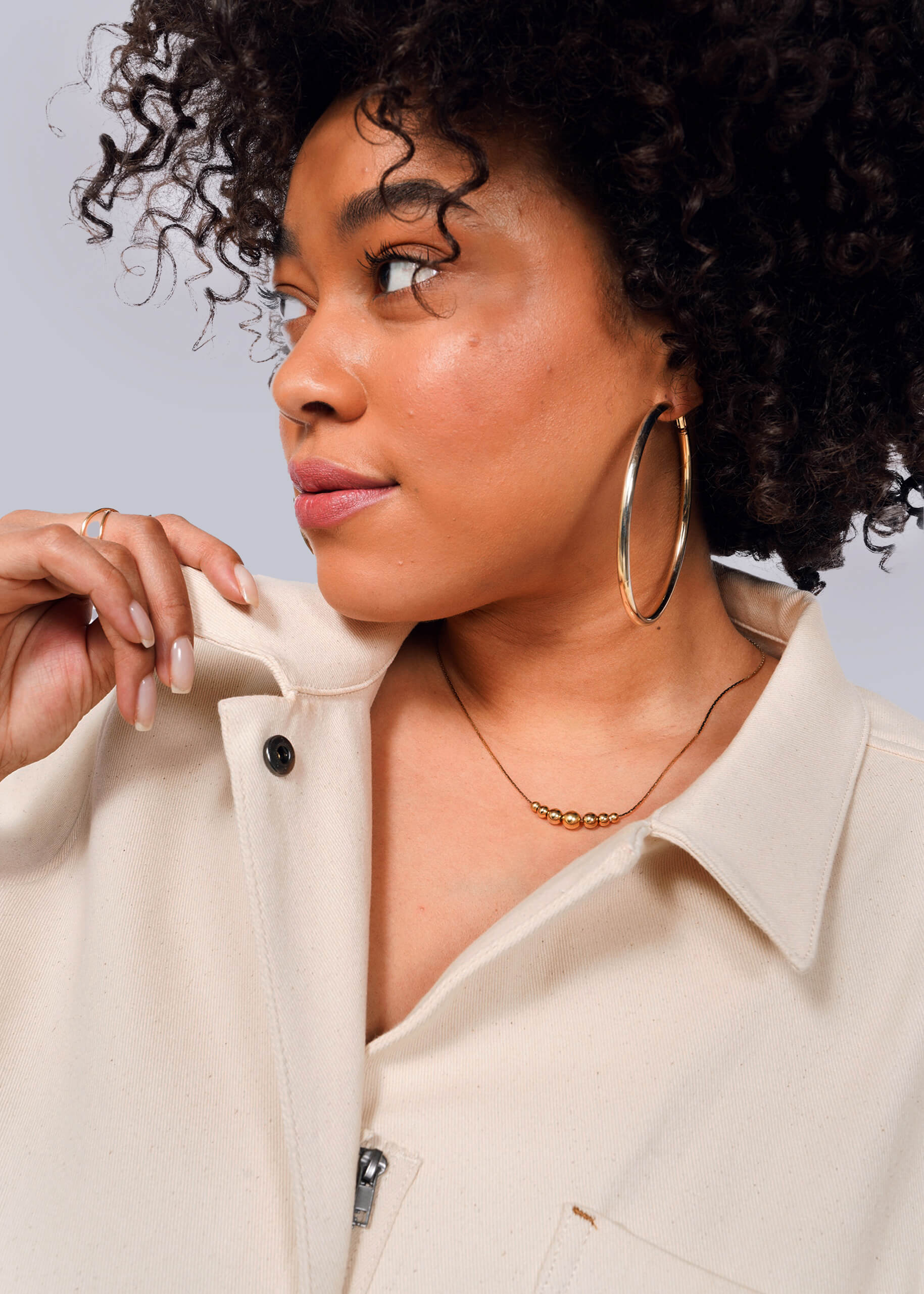 1X=A person with curly hair, large hoop earrings, and a gold necklace is gazing to the side. Theyre holding the collar of a cream-colored jacket over The Essential Denim High Waisted Coverall. The background is plain white.