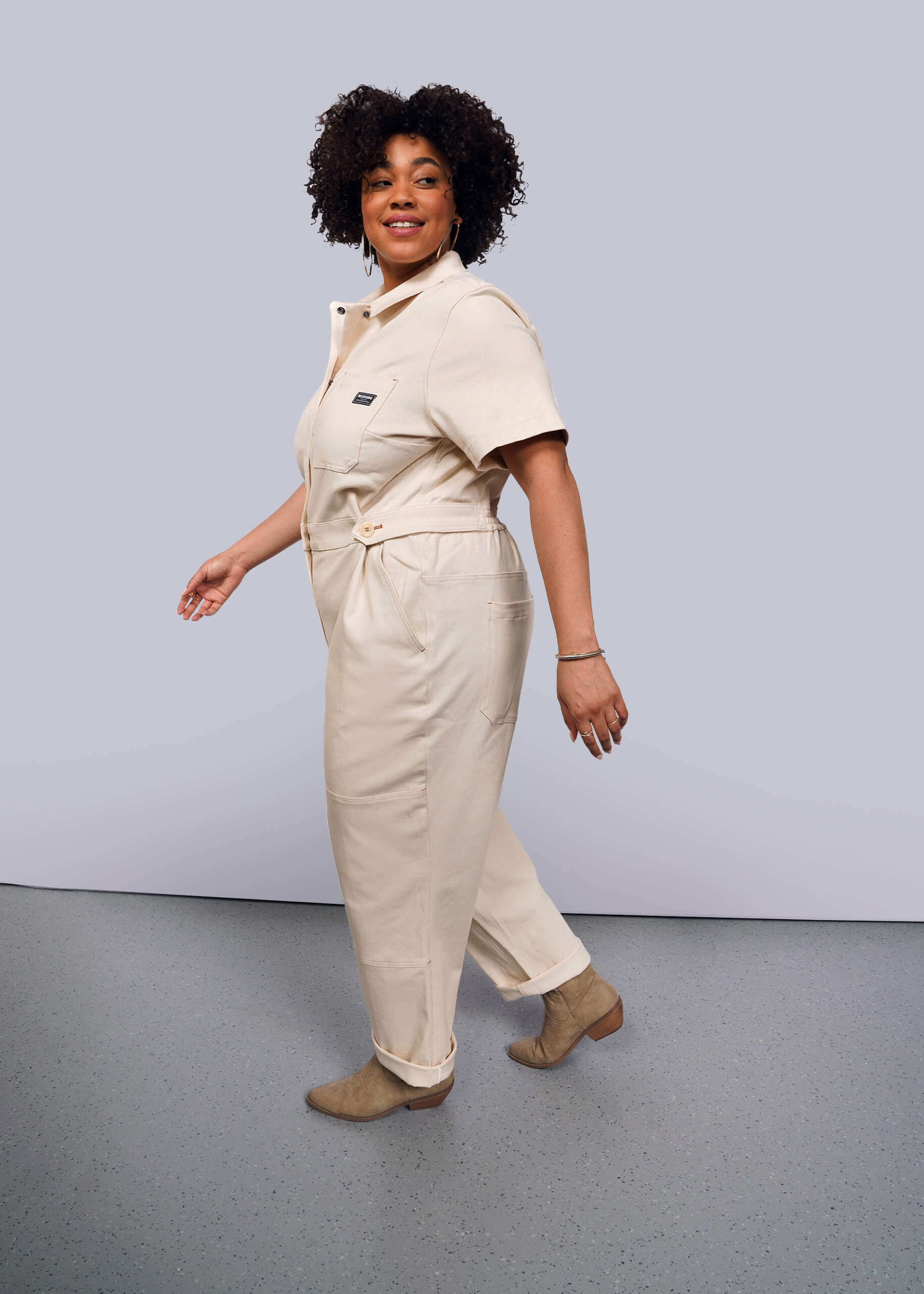 1X=Someone with curly hair is dressed in The Essential Denim High Waisted Coverall featuring deep pockets and tan ankle boots. They stand on a gray floor, slightly turned with one foot forward, smiling against a plain white background.