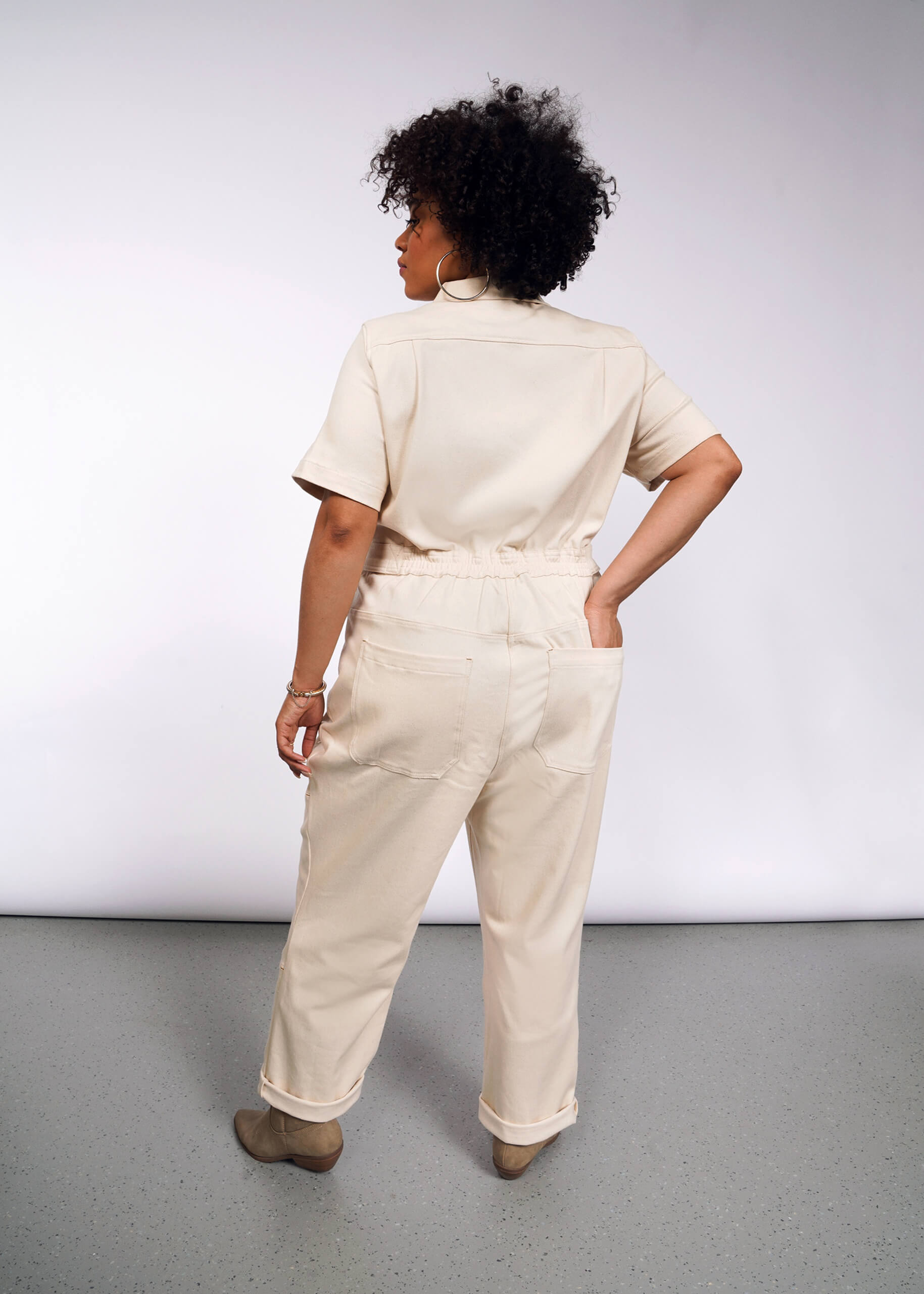 1X=A woman with curly hair stands with her back to the camera, wearing The Essential Denim High Waisted Coverall in beige and matching shoes. She tucks her left hand into one of the deep back pockets against a gray background.