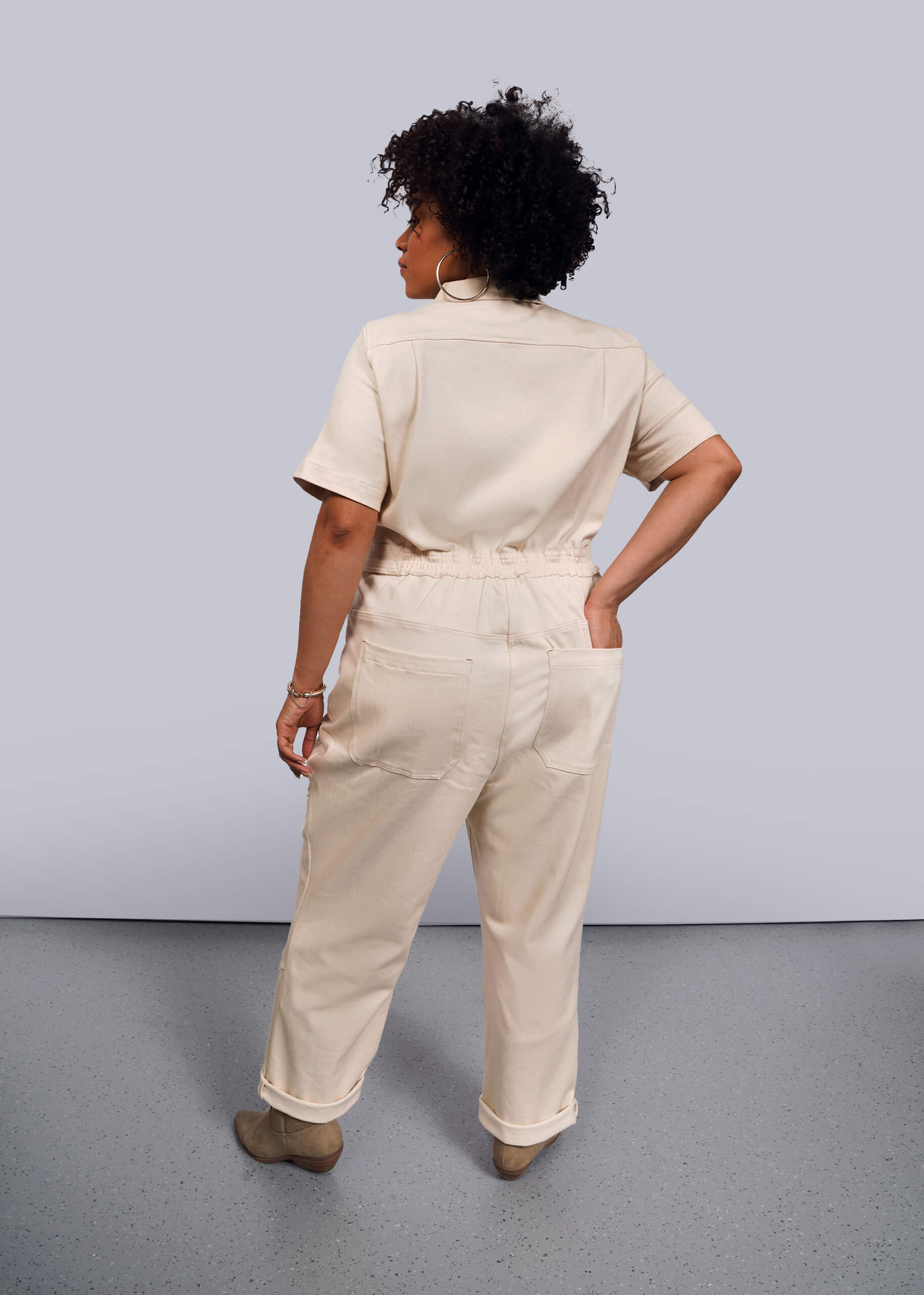1X=A woman with curly hair stands with her back to the camera, wearing The Essential Denim High Waisted Coverall in beige and matching shoes. She tucks her left hand into one of the deep back pockets against a gray background.