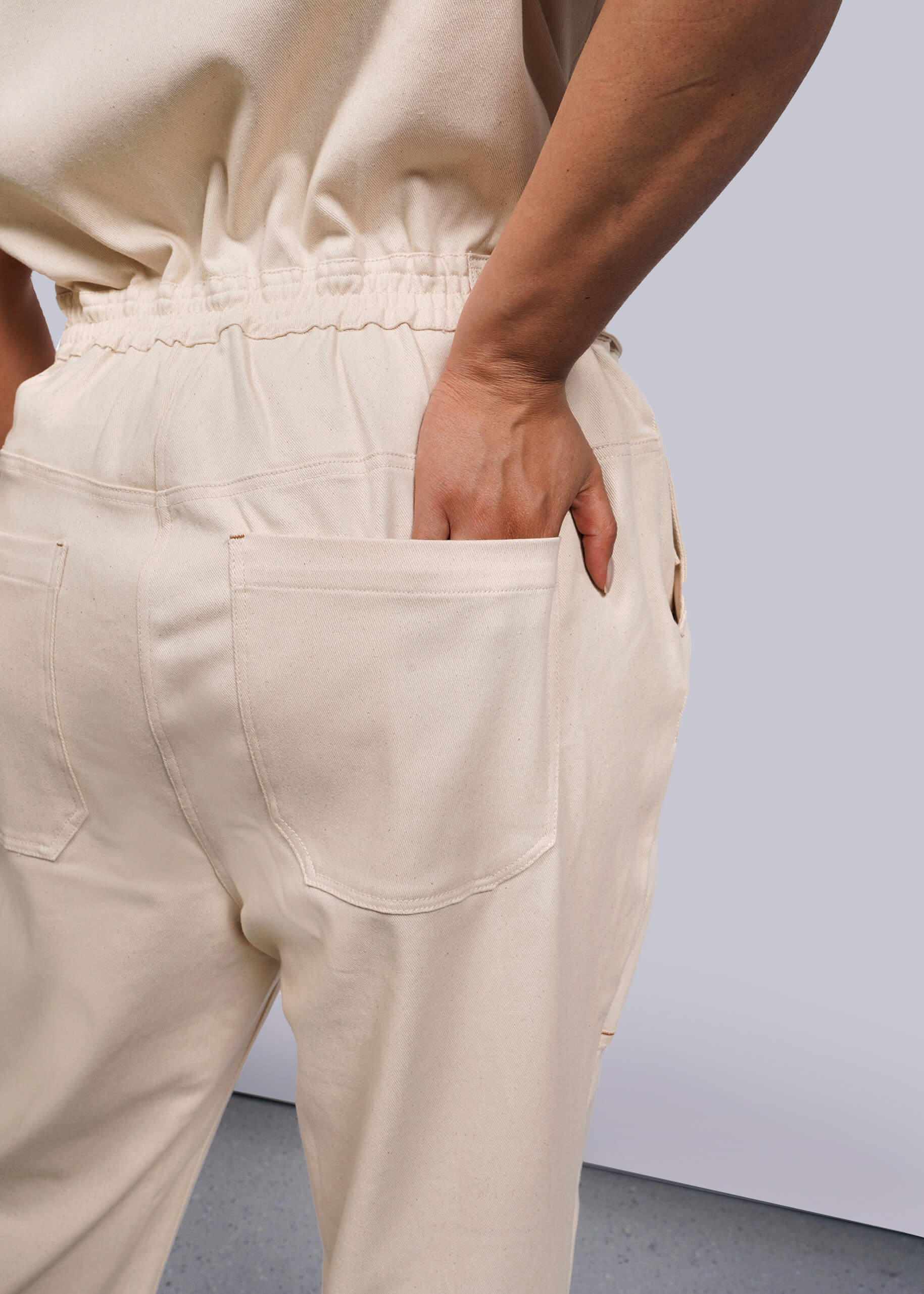 1X=A person is seen from the back wearing The Essential Denim High Waisted Coverall, featuring deep pockets and an elastic waistband. Their left hand rests casually in one pocket, while the light-colored background highlights the simple elegance of this undyed cream-colored garment.