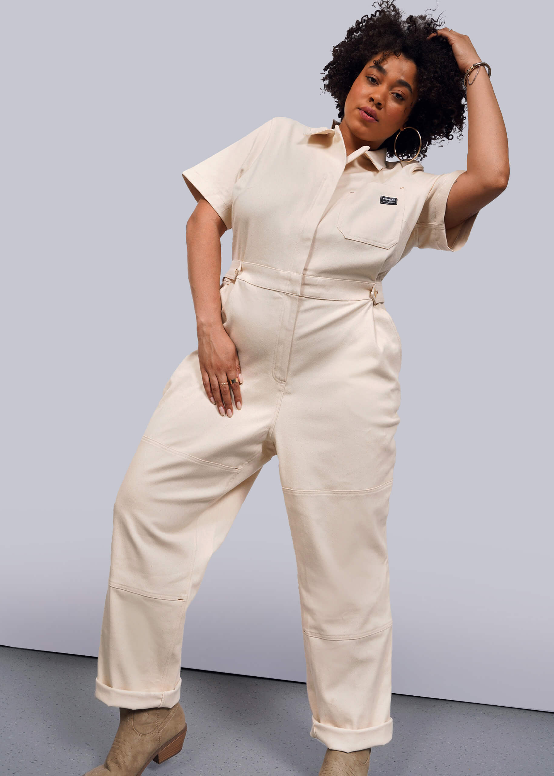 1X= A curly-haired person confidently poses in The Essential Denim High Waisted Coverall, which is cream-colored and features deep pockets, paired with beige boots. They stand on a light gray floor against a white background, with one hand on their head and the other on their hip.