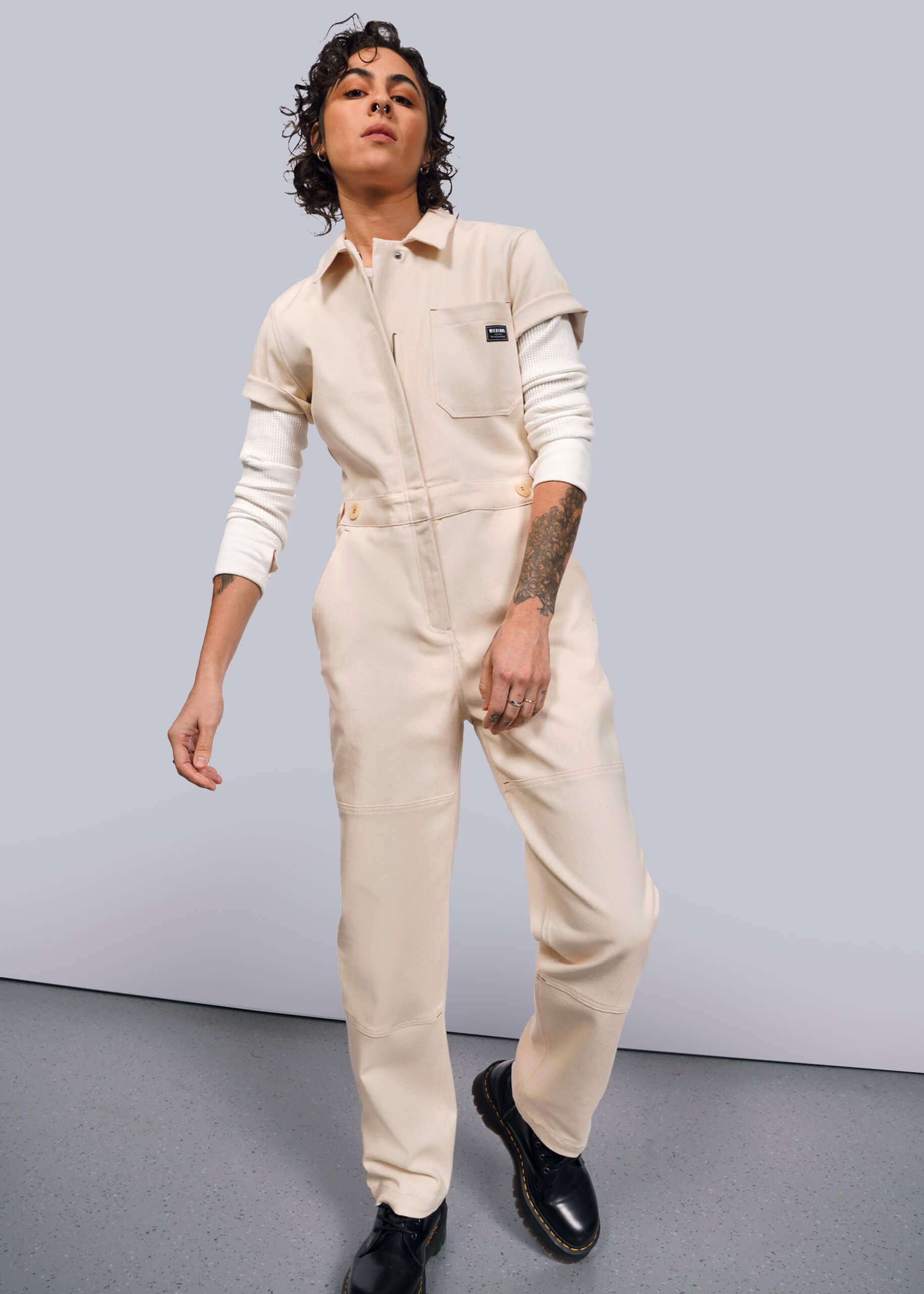 S= A person with curly hair confidently poses in The Essential Denim High Waisted Coverall over a white long-sleeve shirt, showcasing tattoos on one arm, against a plain background.