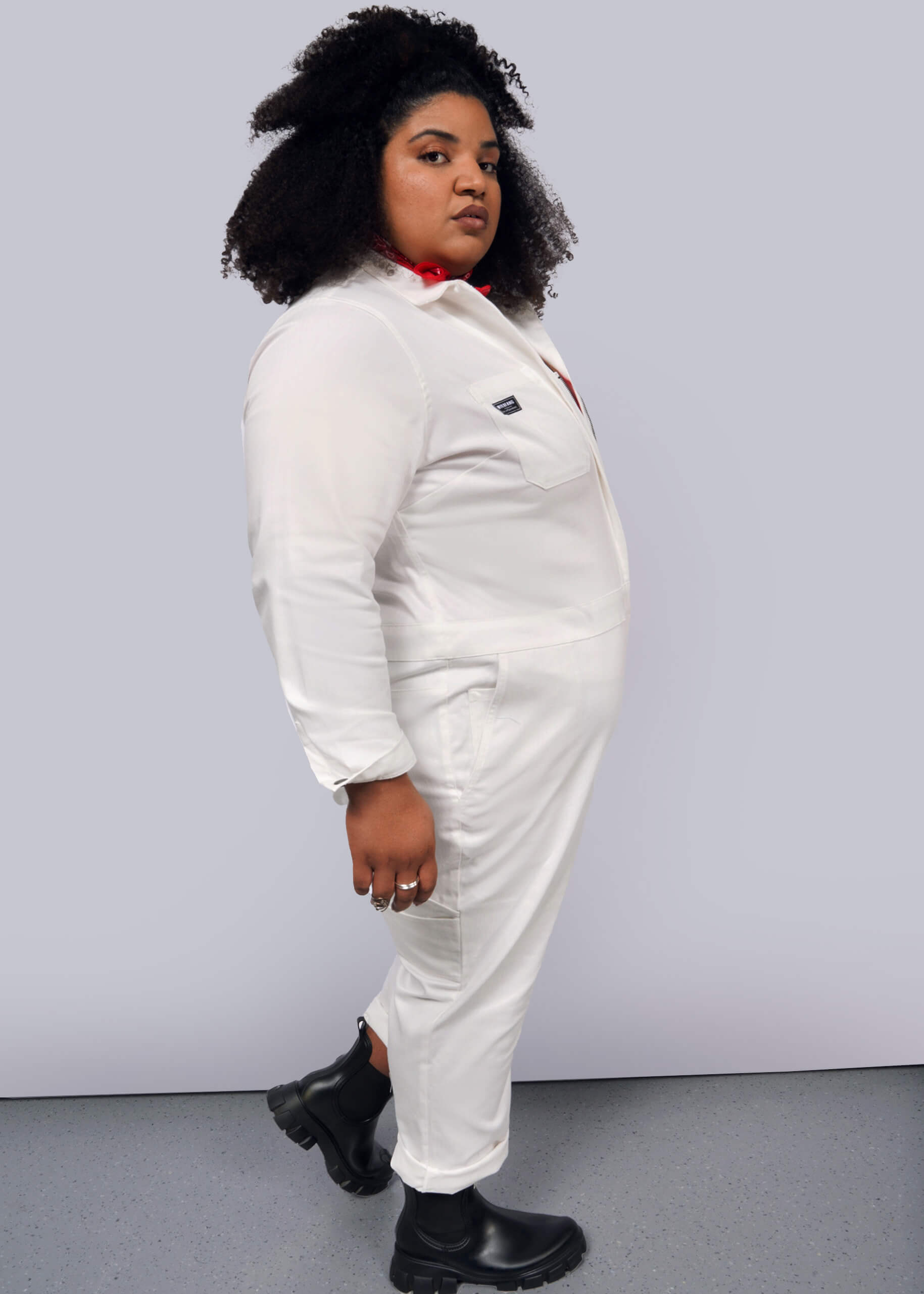 1X=A person in The Essential Long Sleeve Herringbone Coverall, styled with black boots and a red scarf, stands against a gray background. Their curly hair frames their face as they look sideways, adding brightness to the minimalist look.