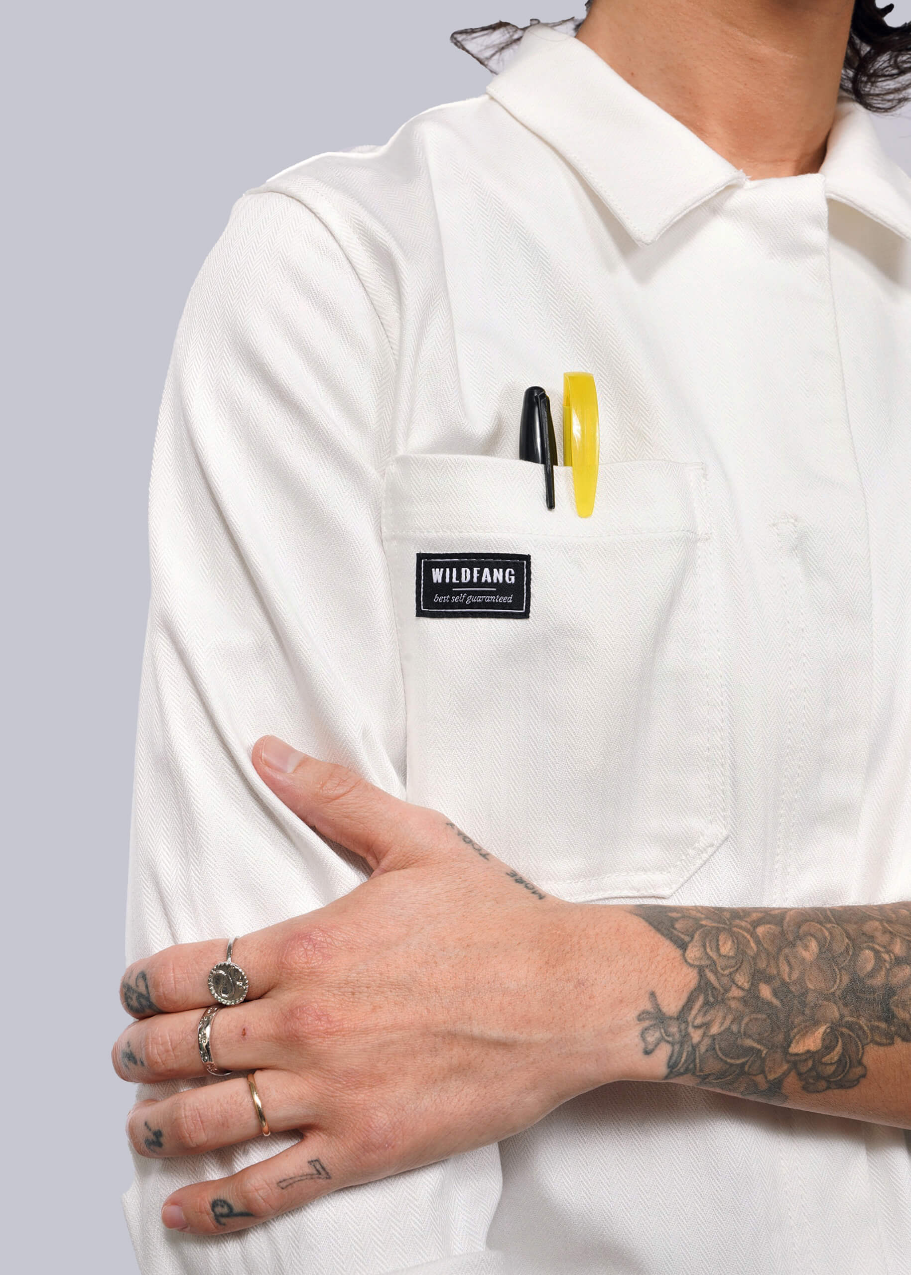 S= In a white Wildfang Essential Long Sleeve Herringbone Coverall, featuring herringbone cotton and 10 pockets, a person stands confidently. Pens peek from one pocket. Tattoos cover an arm, while gleaming rings adorn fingers as they cross arms in front of their body.