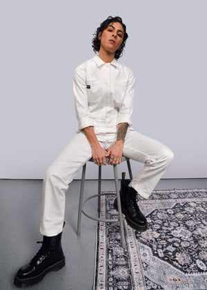 S= A curly-haired person sits on a stool in The Essential Long Sleeve Herringbone Coverall with a full-length zipper and black boots. Their relaxed arms rest on their knees against a plain backdrop, with part of a patterned rug visible on the floor.