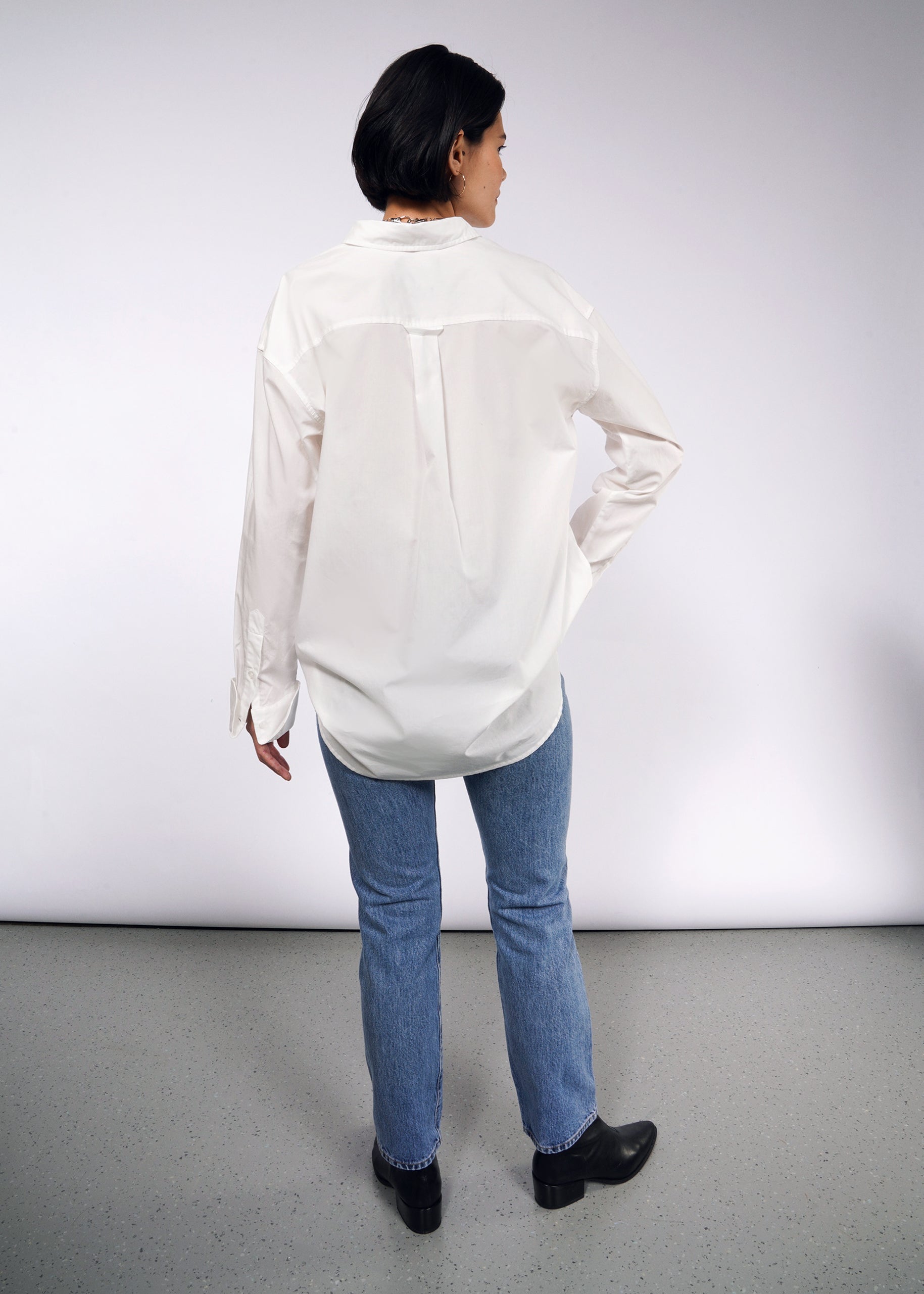 S=With their back to the camera, a person wears The Essential Long Sleeve Oversized Wrap Button Up, paired with blue jeans and black shoes. The background is plain and light-colored.