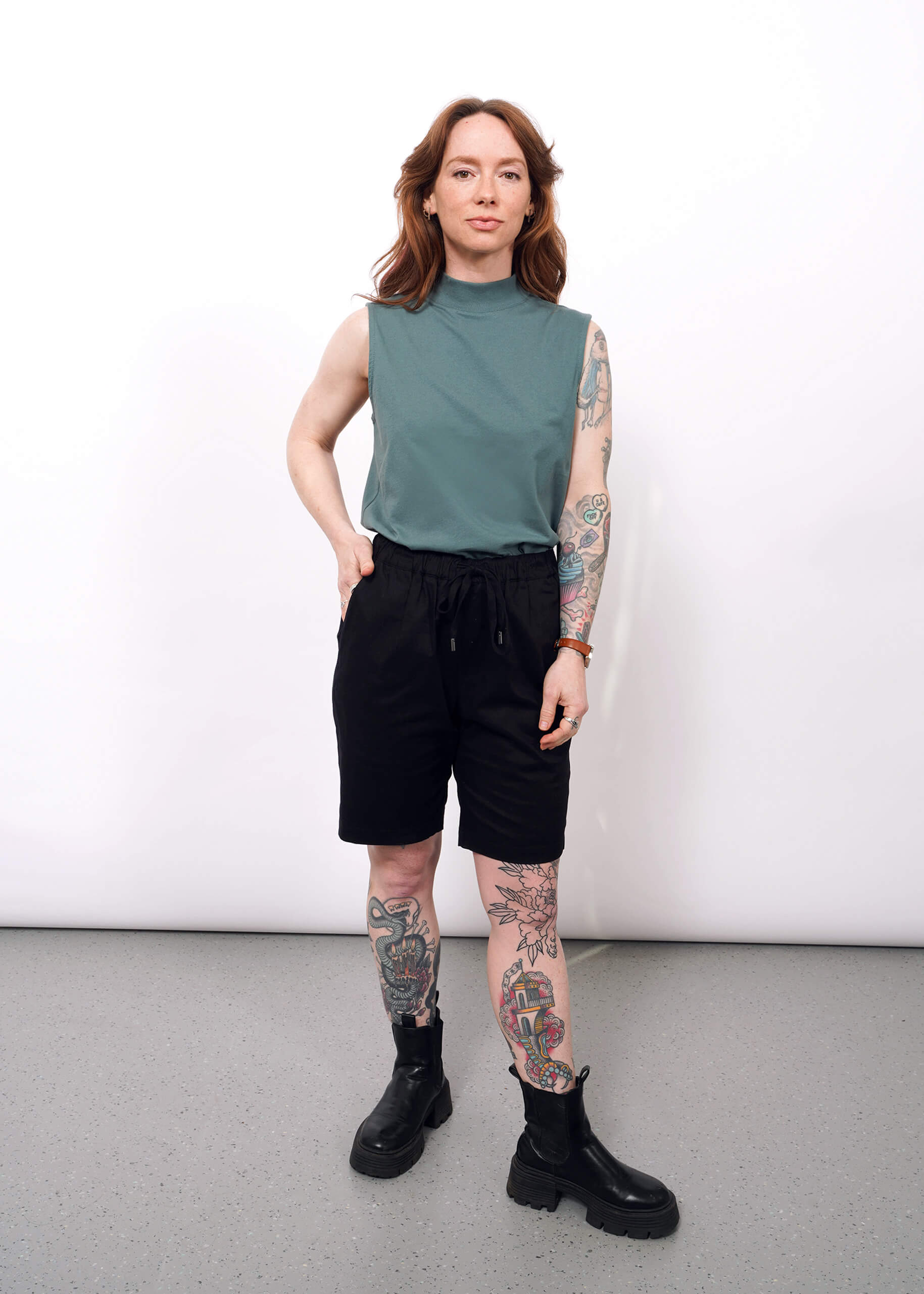 S=A person with long, wavy brown hair and visible tattoos stands confidently against a plain white wall, styled in The Essential Mock Neck Tank in green with black shorts and boots—a perfect layering piece for any casual outfit.