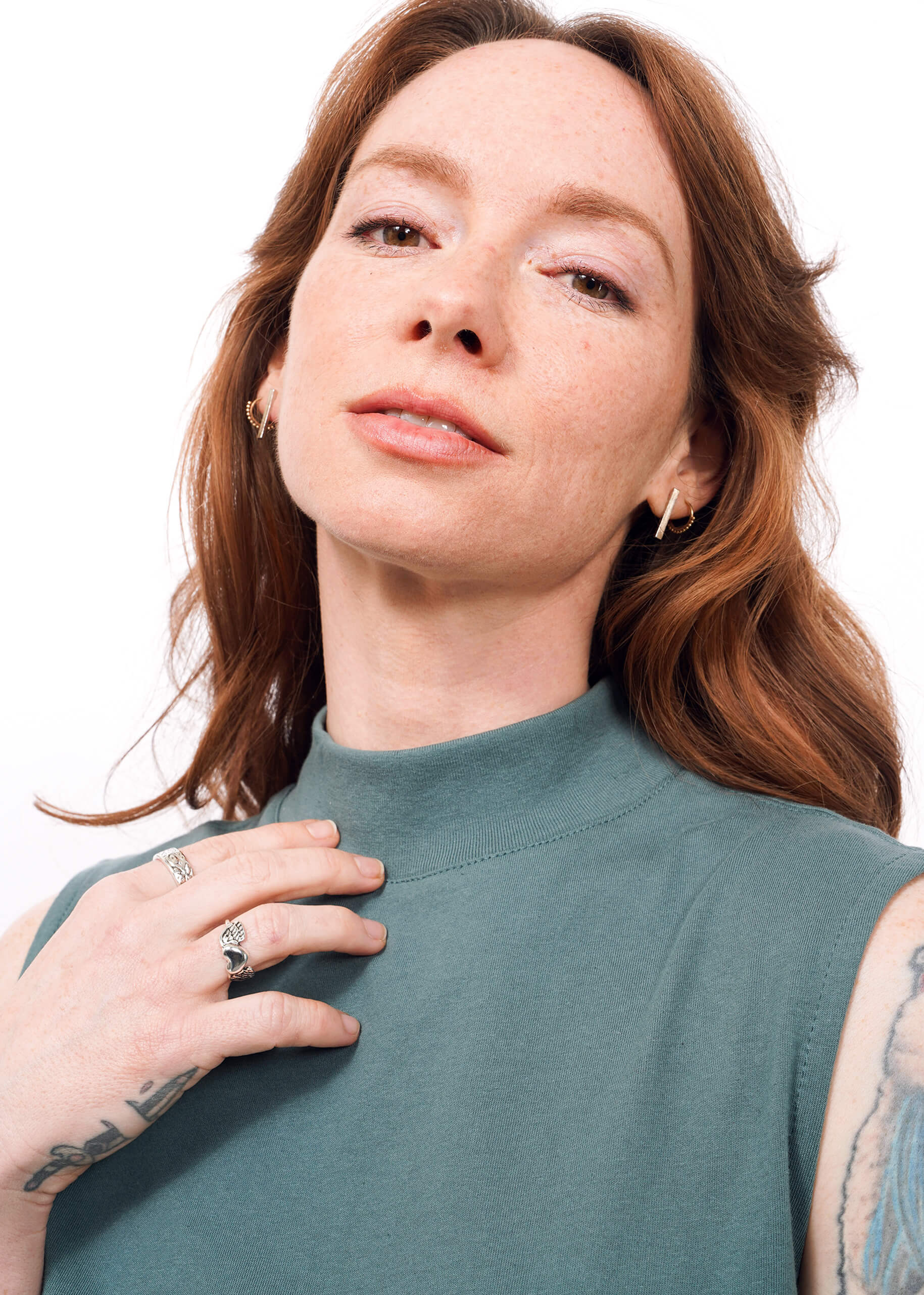 S= A woman with wavy red hair and visible hand tattoos confidently gazes at the camera, wearing The Essential Mock Neck Tank in green. She has light freckles and subtle makeup, with her hand resting gently on her chest.