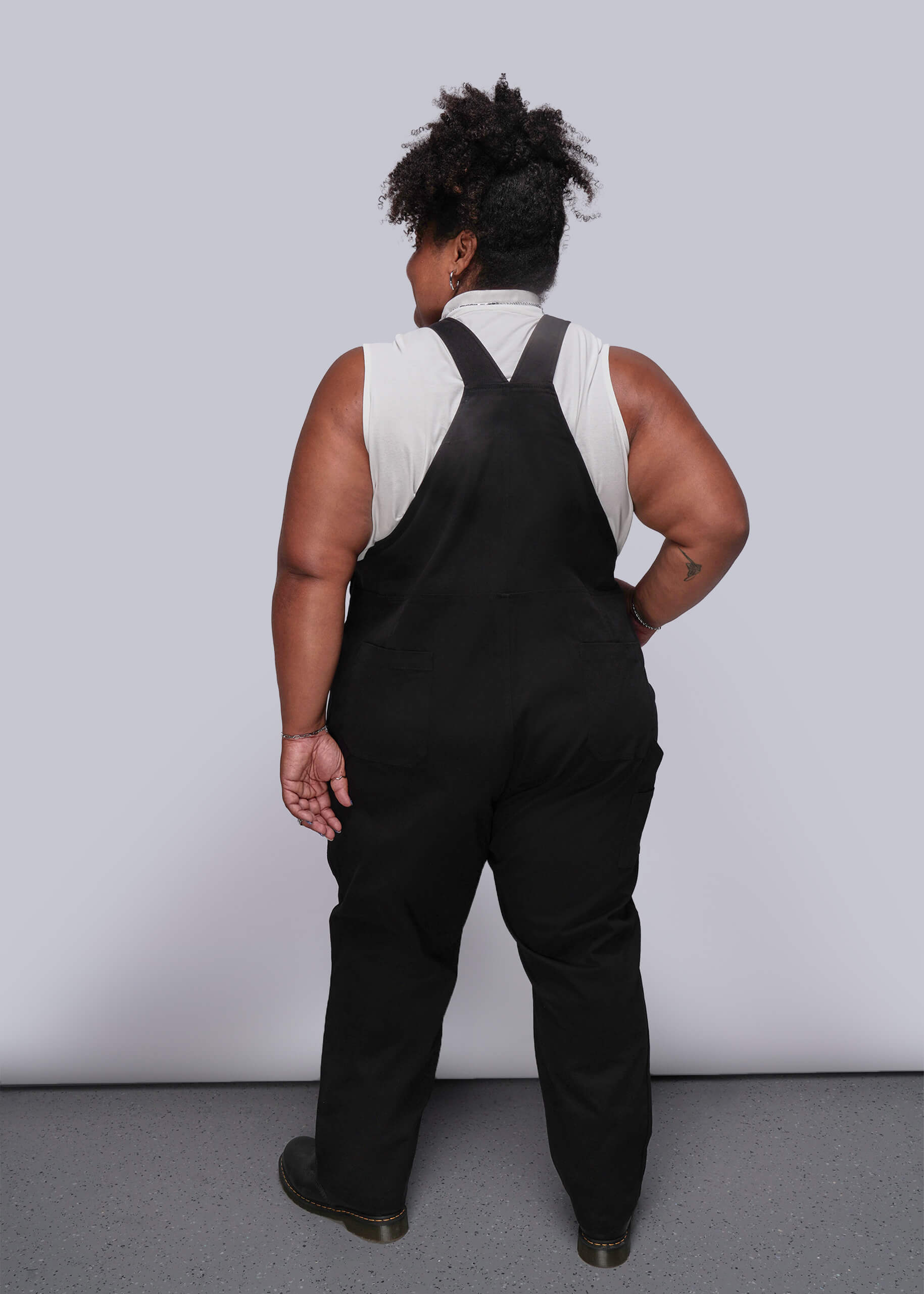 1X=Person wearing black overalls with a white shirt underneath against a plain background