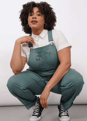 1X=A person with curly hair wearing The Essential Overall—teal, with adjustable straps and deep pockets—over a white button up shirt and paired with black Converse sneakers, squats and looks at camera against a plain white background.