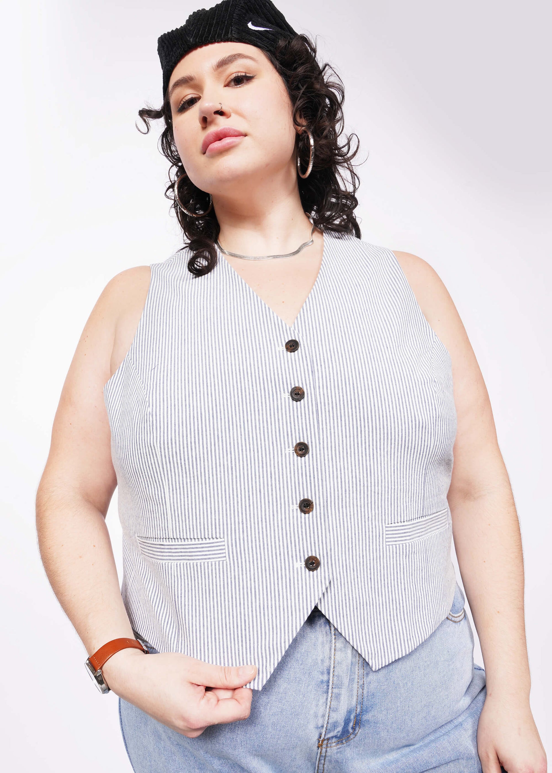 1X= Person with short curly black hair looks at camera wearing seersucker blue and white vest and jeans.