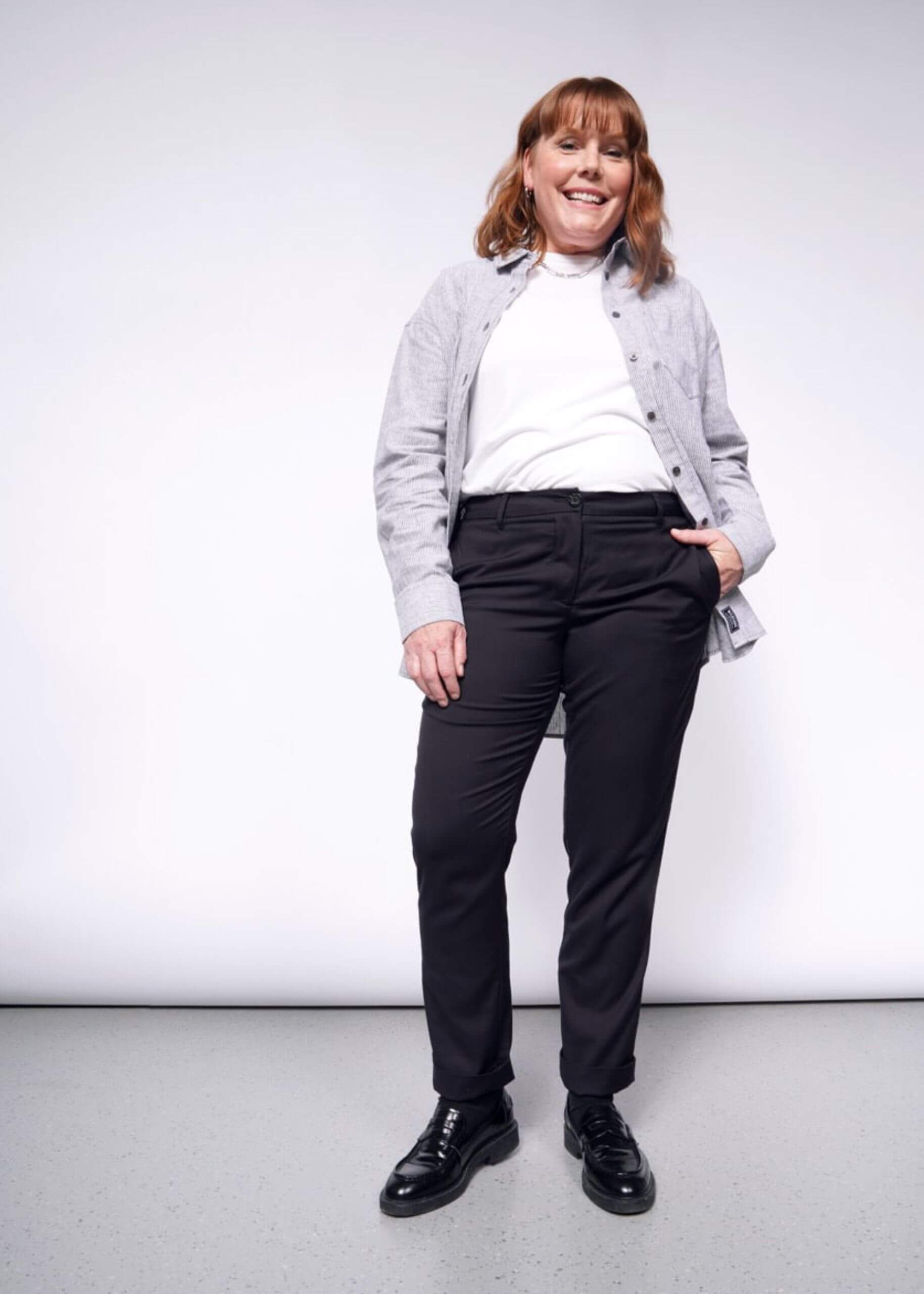 S=A person with long brown hair smiles in a white shirt and gray jacket, paired with The Essential Trouser in dark color and black shoes. They stand against a light gray background, hands in pockets.