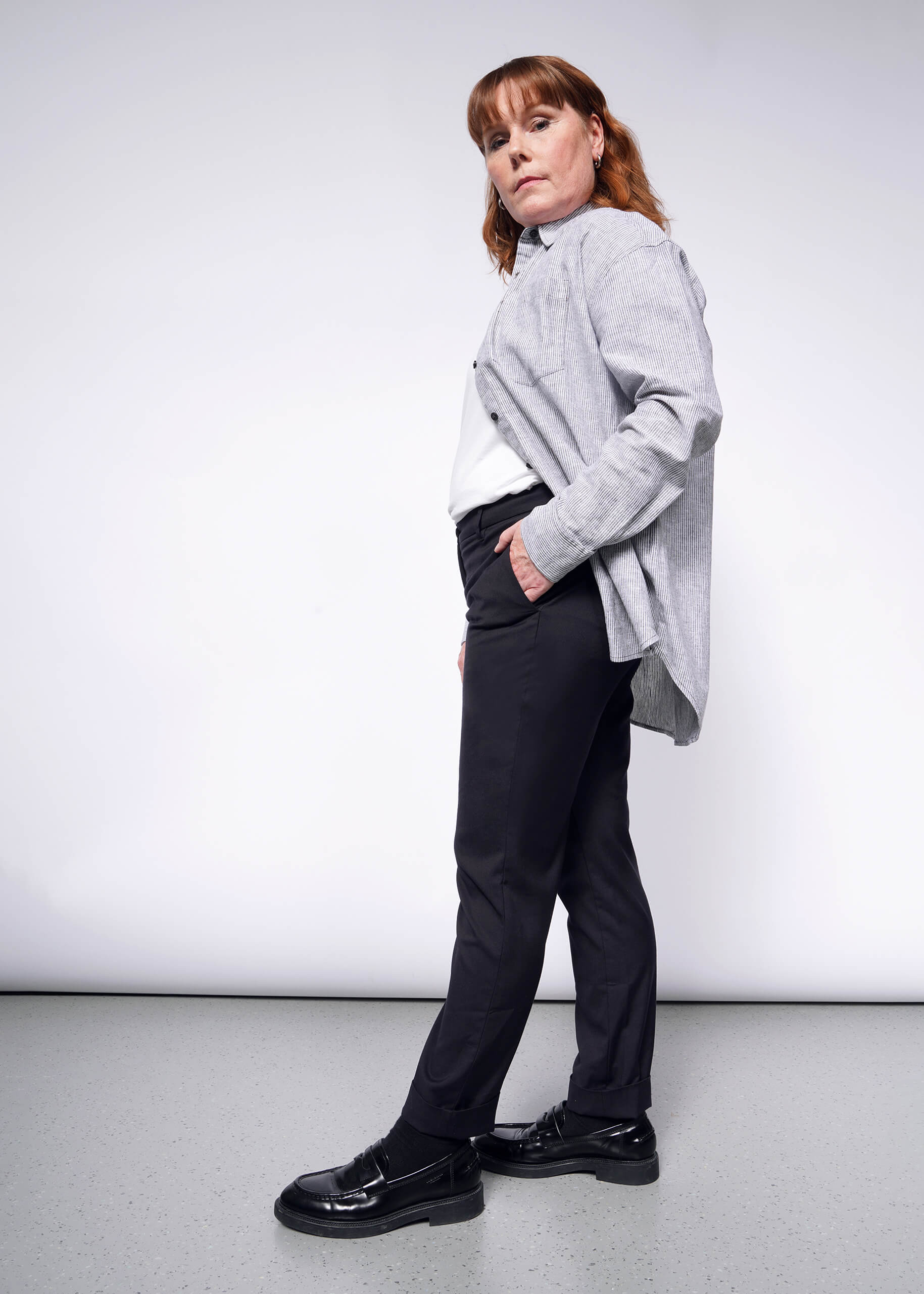S=A person with medium-length red hair confidently wears a gray button-up shirt with convertible cuffs, paired with The Essential Trouser in black and matching shoes. They pose casually against a plain white background, one hand tucked into their pocket.