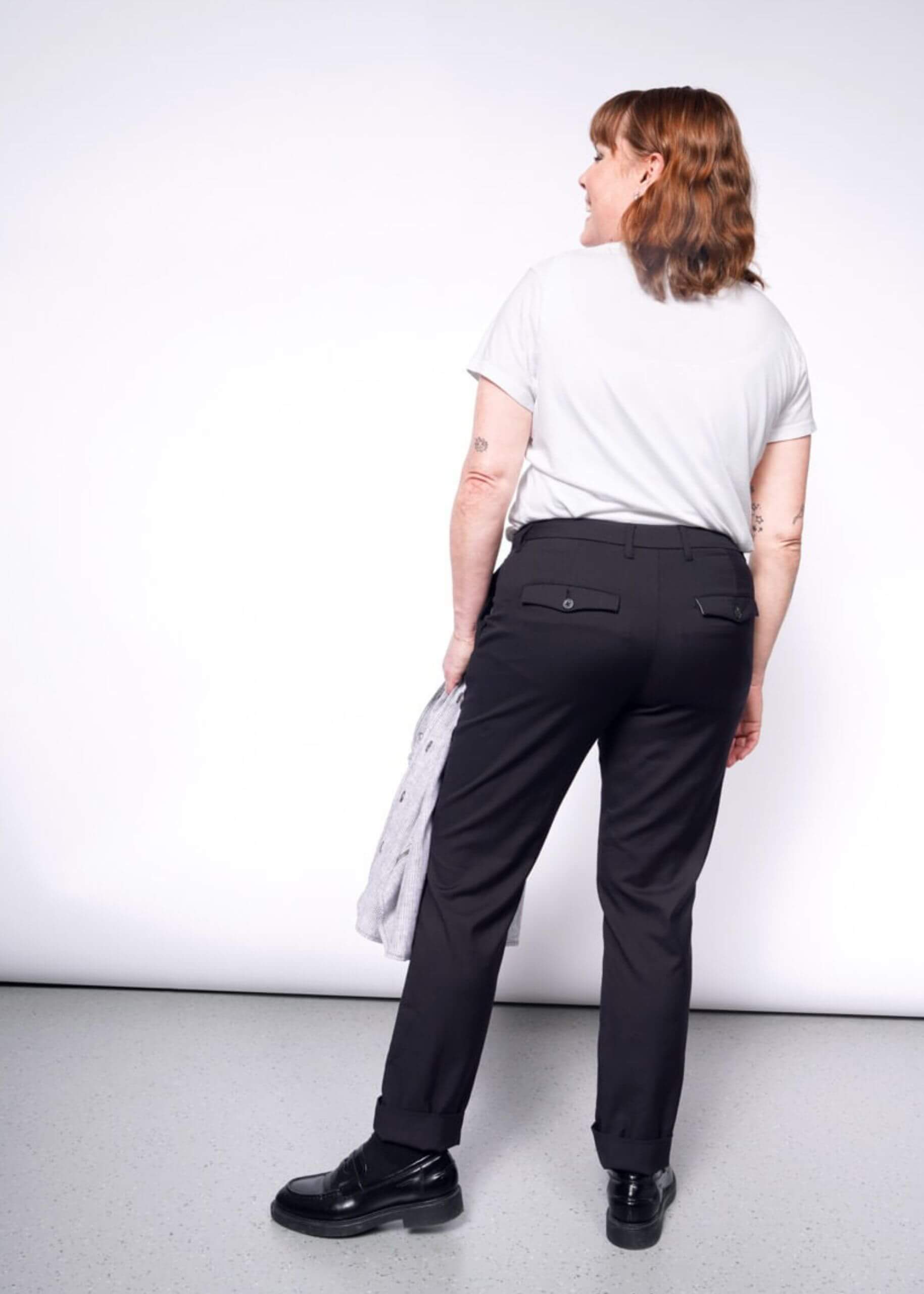 S=A person with shoulder-length hair stands facing away on a plain white background, wearing a white t-shirt and The Essential Trouser in a relaxed mid-rise fit, paired with black shoes and holding a light-colored sweater.