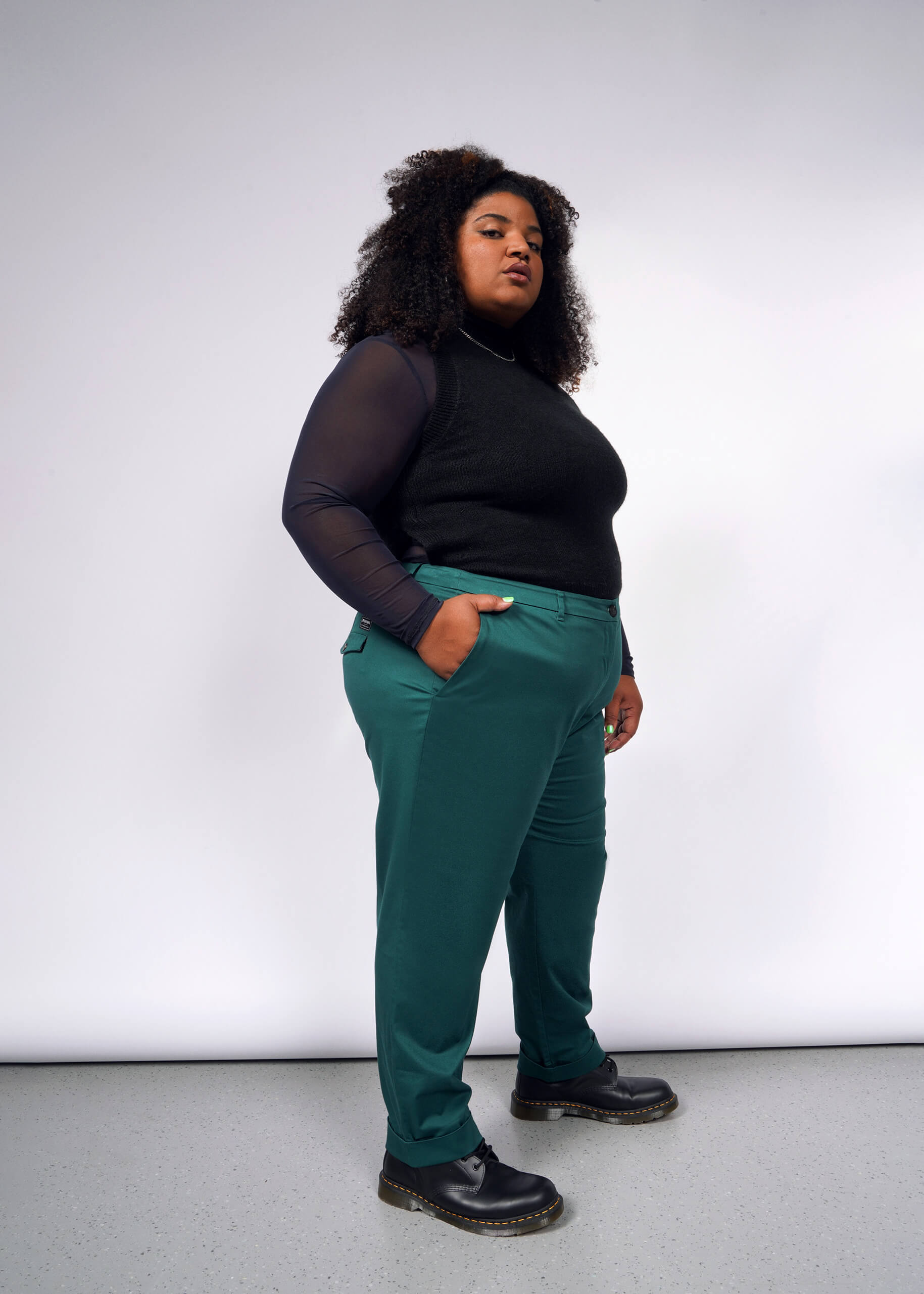 1X=A person stands confidently against a white background, wearing a black top with sheer sleeves, The Essential Trouser in teal, and black boots. They have curly hair and their hands in their pockets.