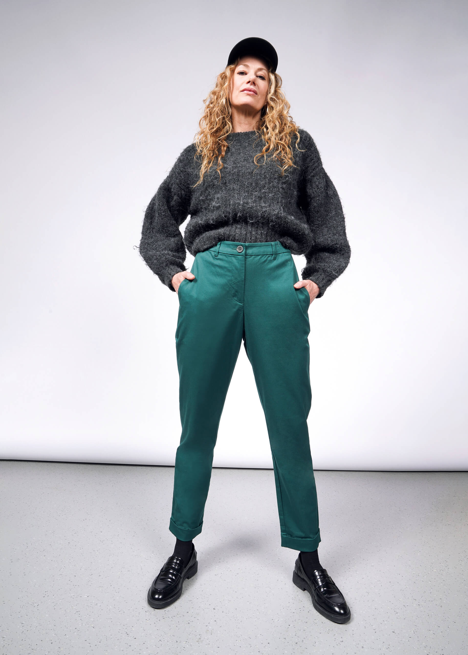 S= A person with long curly hair is wearing a black cap, gray sweater, and The Essential Trouser in green, paired with black shoes. They stand confidently with hands in pockets against a plain background, showcasing their flair for wardrobe staples.