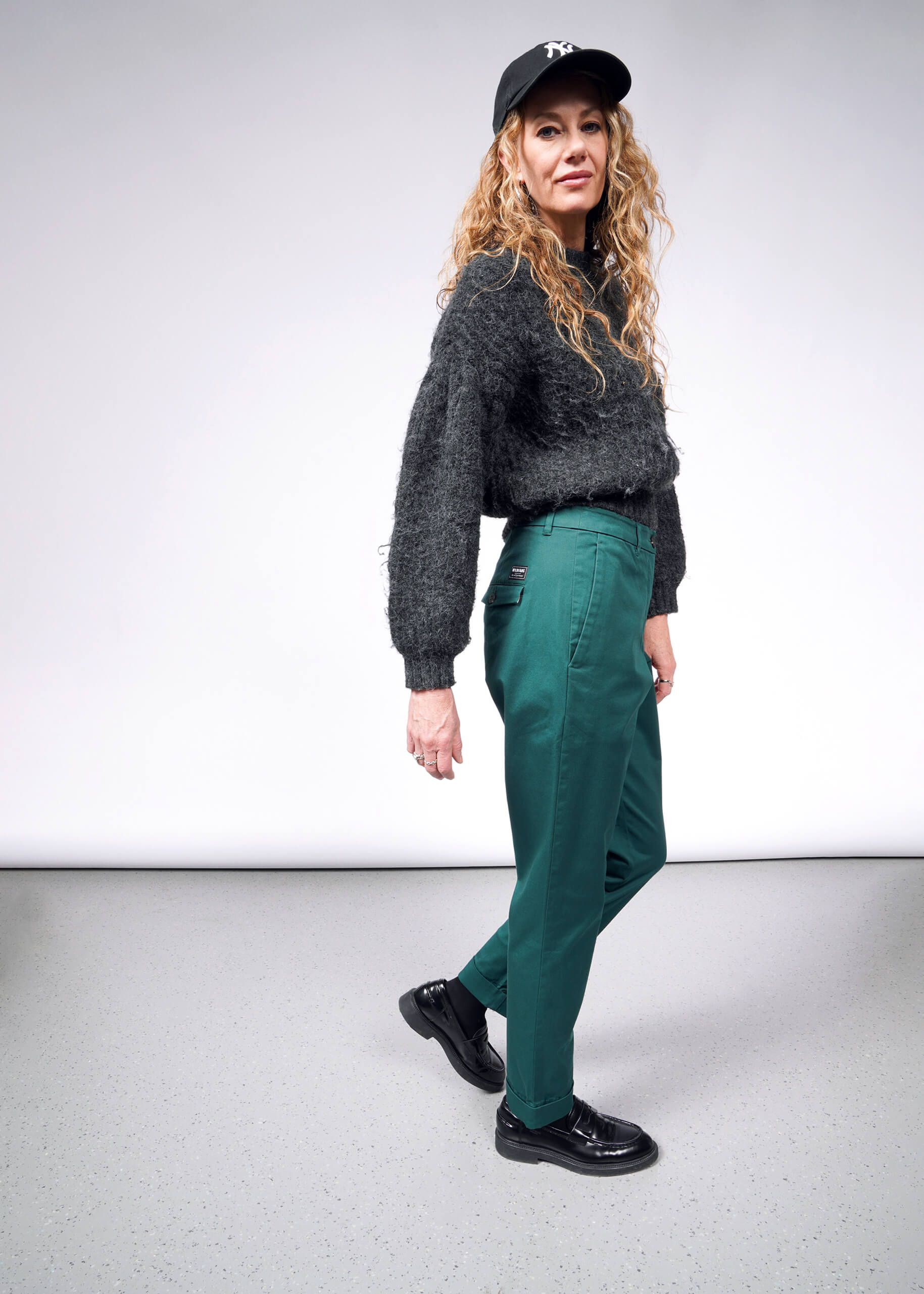 S=A woman with long curly hair wears a dark cap, fuzzy sweater, and The Essential Trouser in green. She poses confidently in shiny black shoes against a plain white and gray backdrop.
