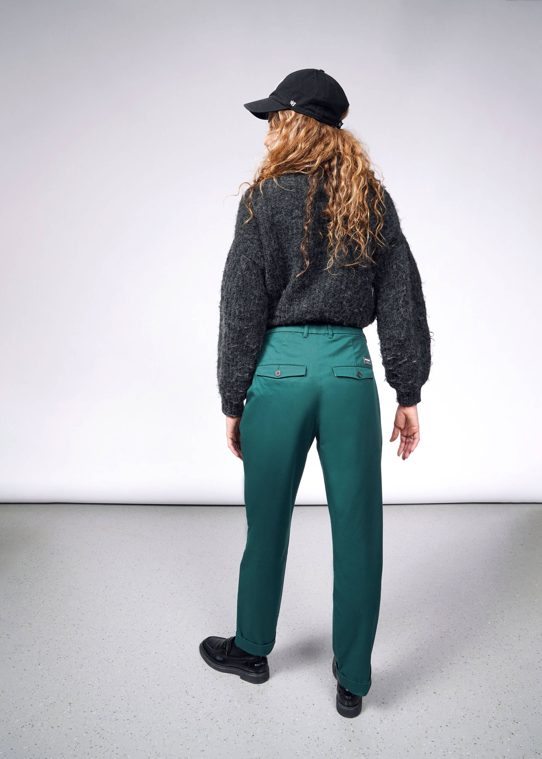 S= A person with long curly hair stands facing away, wearing a black cap, dark gray sweater, The Essential Trouser in green, and black shoes on a light gray floor against a plain background.