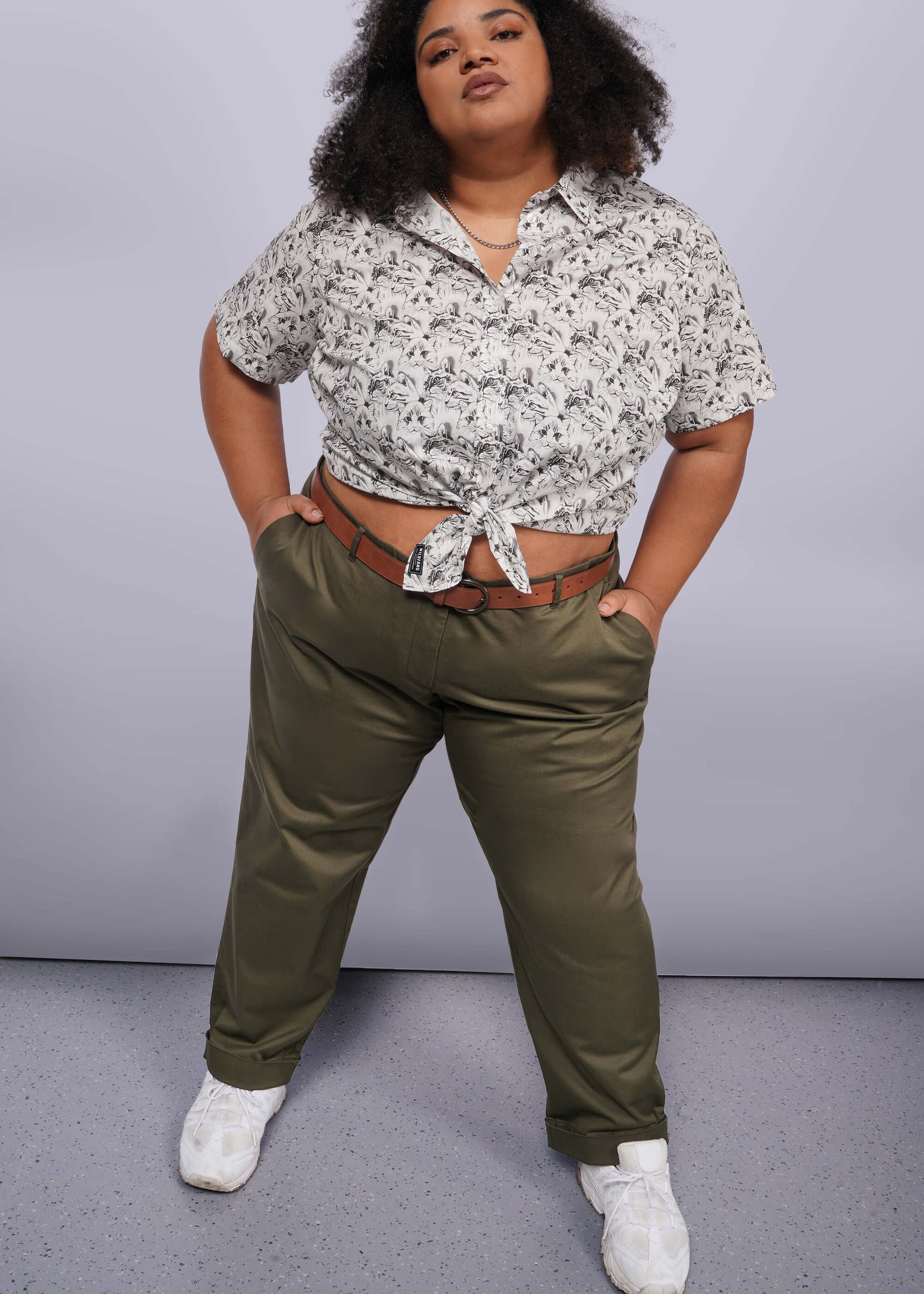 1X=A curly-haired person confidently stands with hands in pockets, wearing a patterned short-sleeve shirt tied at the waist. They pair The Essential Trouser in olive green, a wardrobe staple, with a brown belt and white sneakers against a gray and white gradient background.