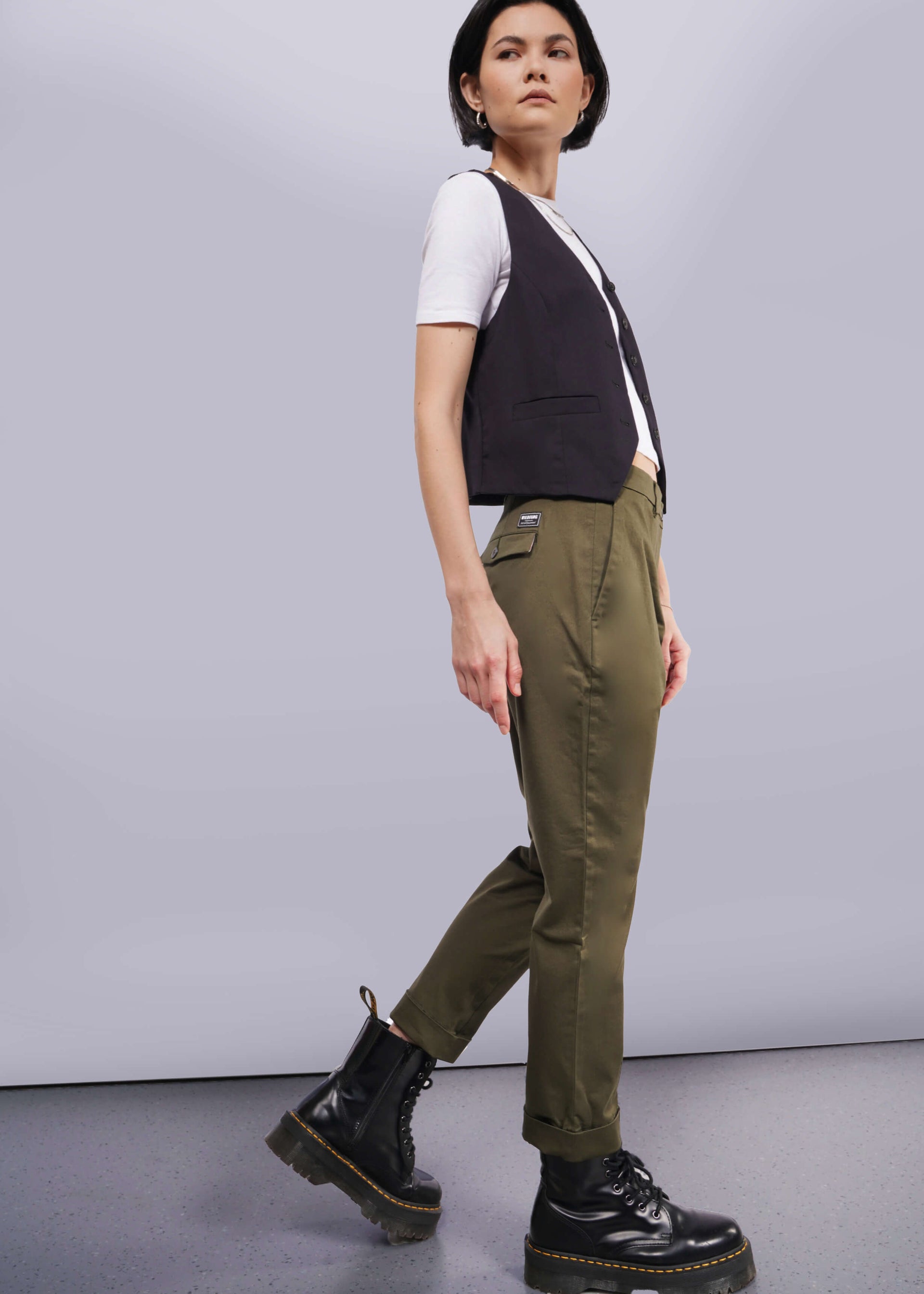 S= Against a plain backdrop, a person sports The Essential Trouser in olive green, paired with a white t-shirt, black vest, and black boots. They have short dark hair and glance sideways.