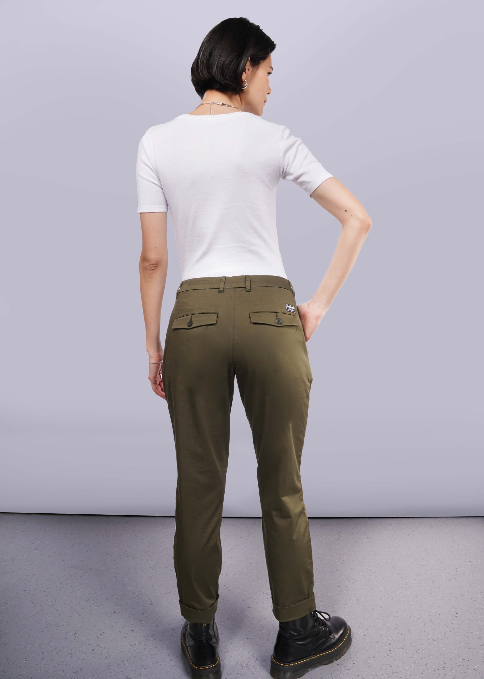 S=Rear view of a person with short dark hair against a white background, wearing a wardrobe staple: a white short-sleeved shirt, The Essential Trouser in classic olive green, and black shoes. They face away with one hand on their hip.