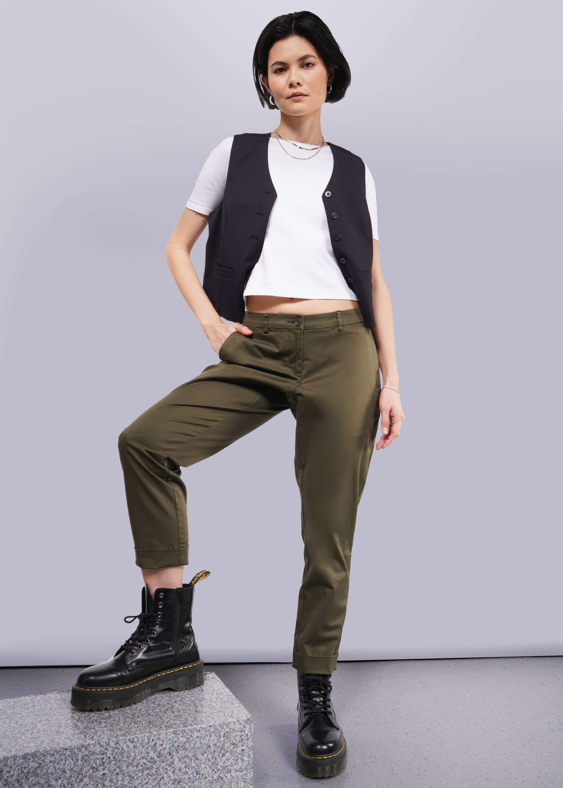 S=A person with short dark hair poses confidently in a white t-shirt, a black vest, and The Essential Trouser in olive green. These wardrobe staples pair seamlessly with their black boots as they stand with one hand in their pocket against a neutral background.