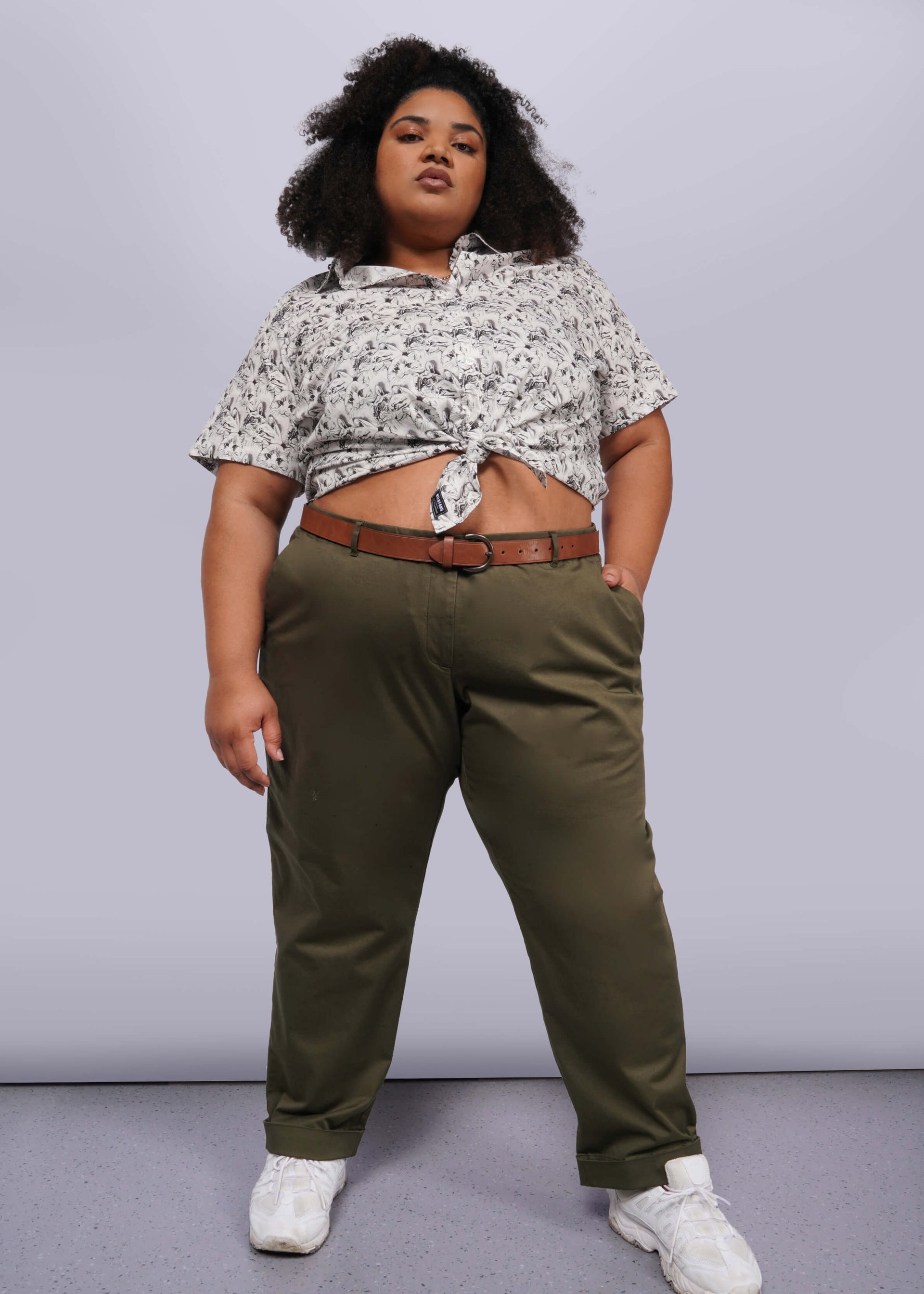 1X= A person stands confidently with one hand in their pocket, featuring a knotted patterned shirt, The Essential Trouser, and white sneakers. The plain background accentuates their bold stance and fashion-forward style, highlighting how these trousers are the ultimate wardrobe staple.