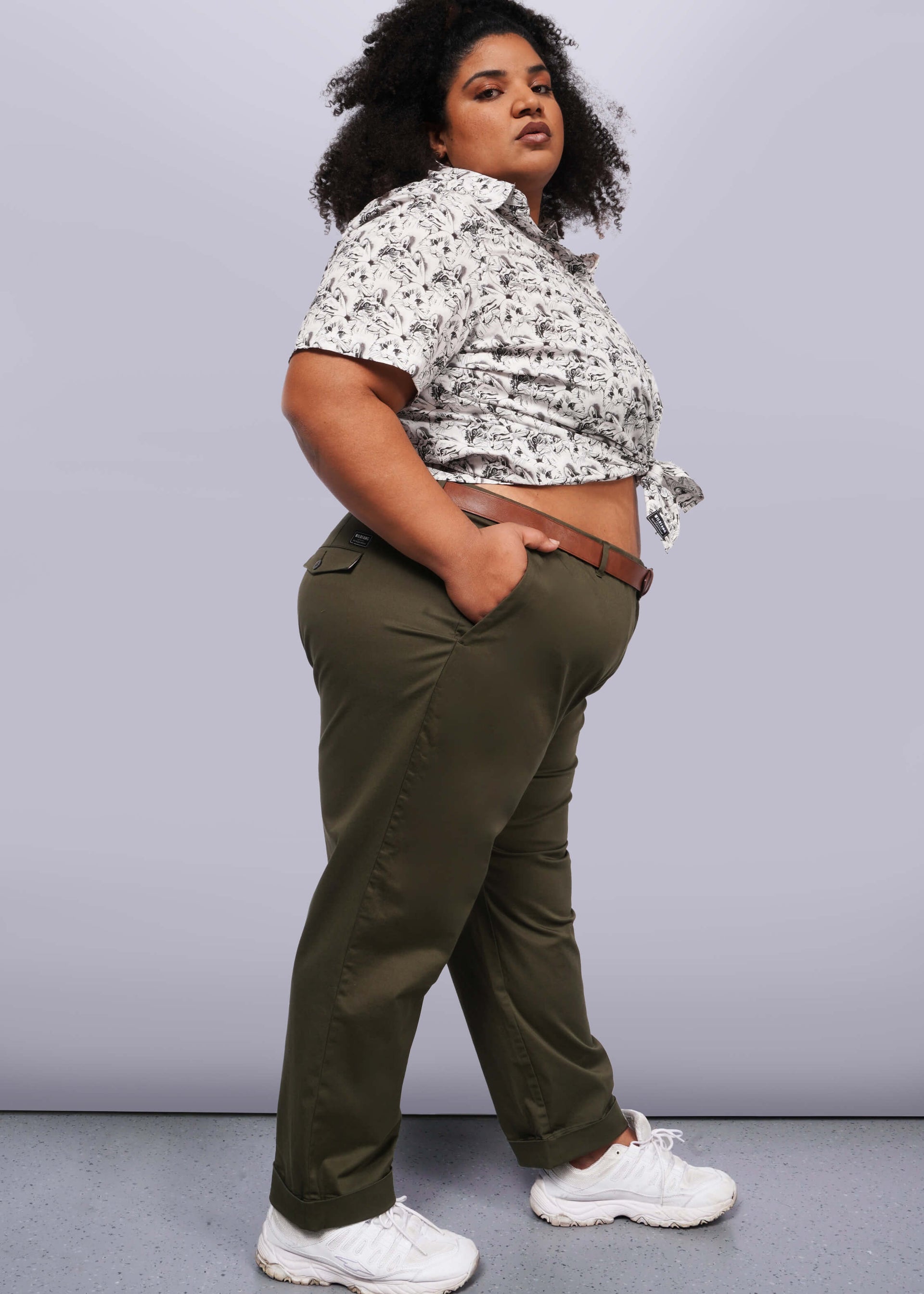 1X=A person wearing a patterned short-sleeve shirt, The Essential Trouser, and white sneakers stands with one hand in their pocket. They face sideways, confidently looking at the camera against a plain grey backdrop—a true wardrobe staple capturing effortless style.