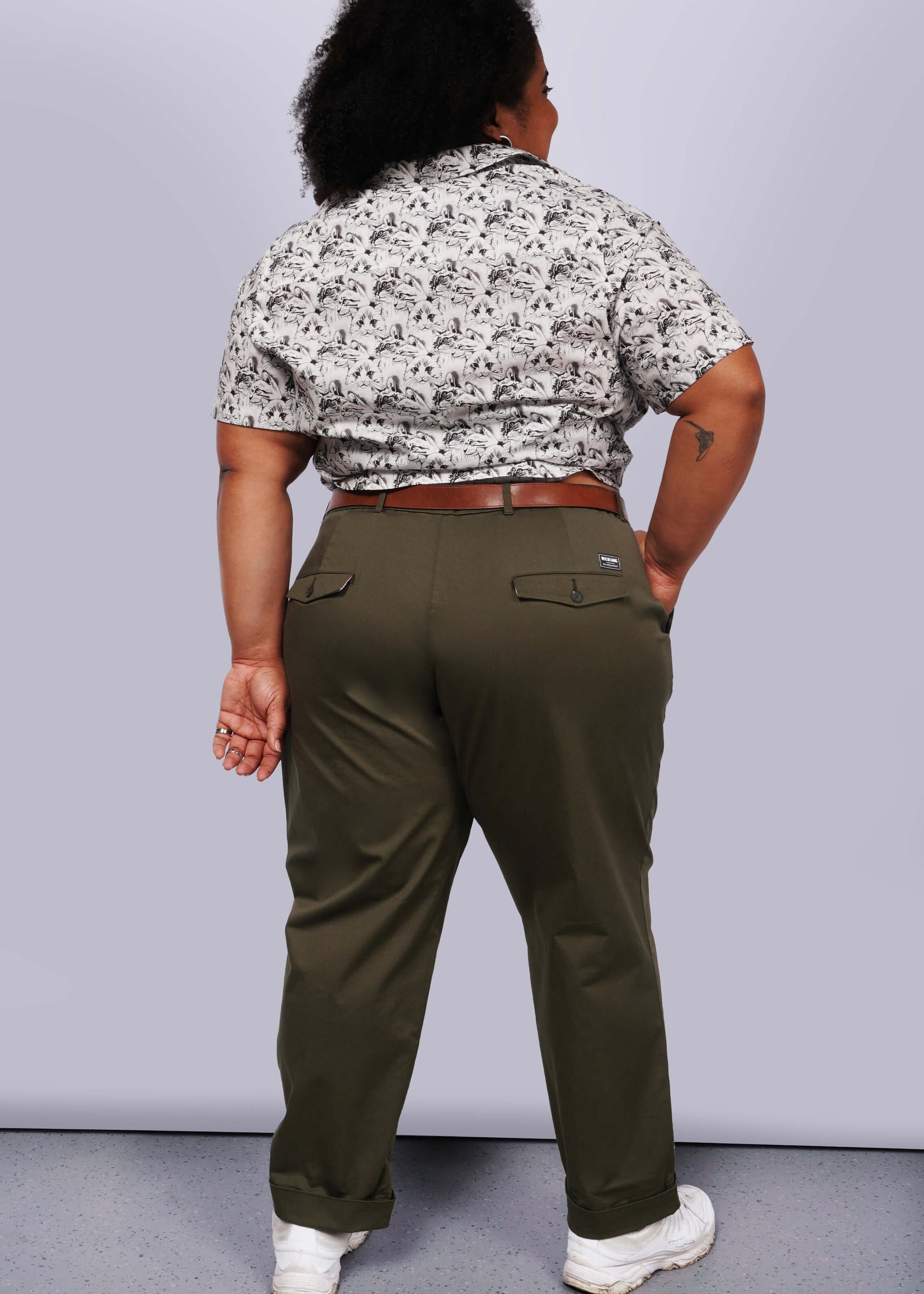 1X= Against a white backdrop, a person faces away, wearing a patterned shirt with convertible cuffs and The Essential Trouser in olive green, belt-cinched as a wardrobe classic. Their feet are adorned with white sneakers, and their right forearm features an intriguing tattoo.