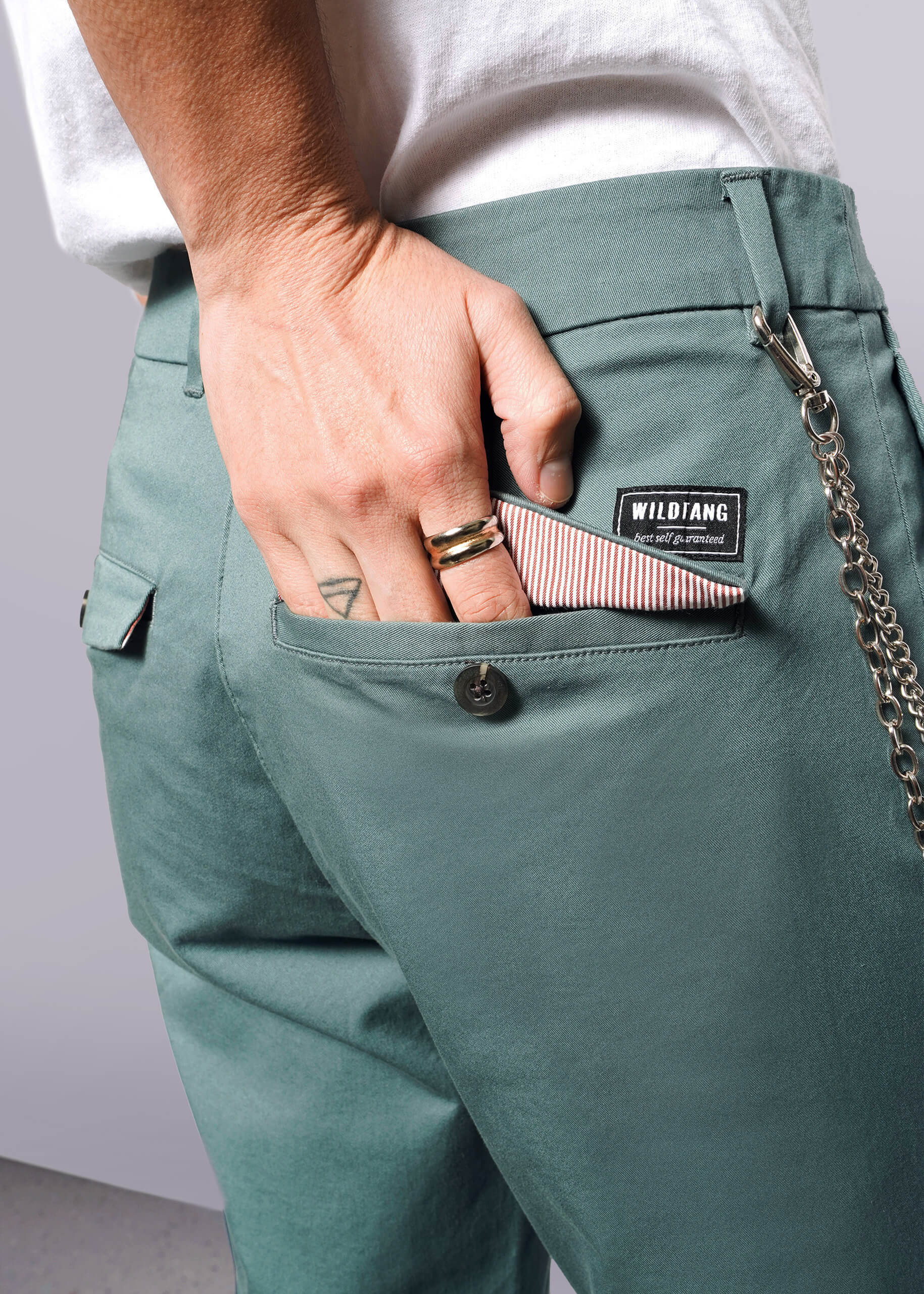 S=Wearing The Essential Trouser with a WILDIANG patch and relaxed mid rise fit, a person puts a striped wallet in their back pocket. Rings decorate their fingers, and a chain dangles from the classic trouser pocket.