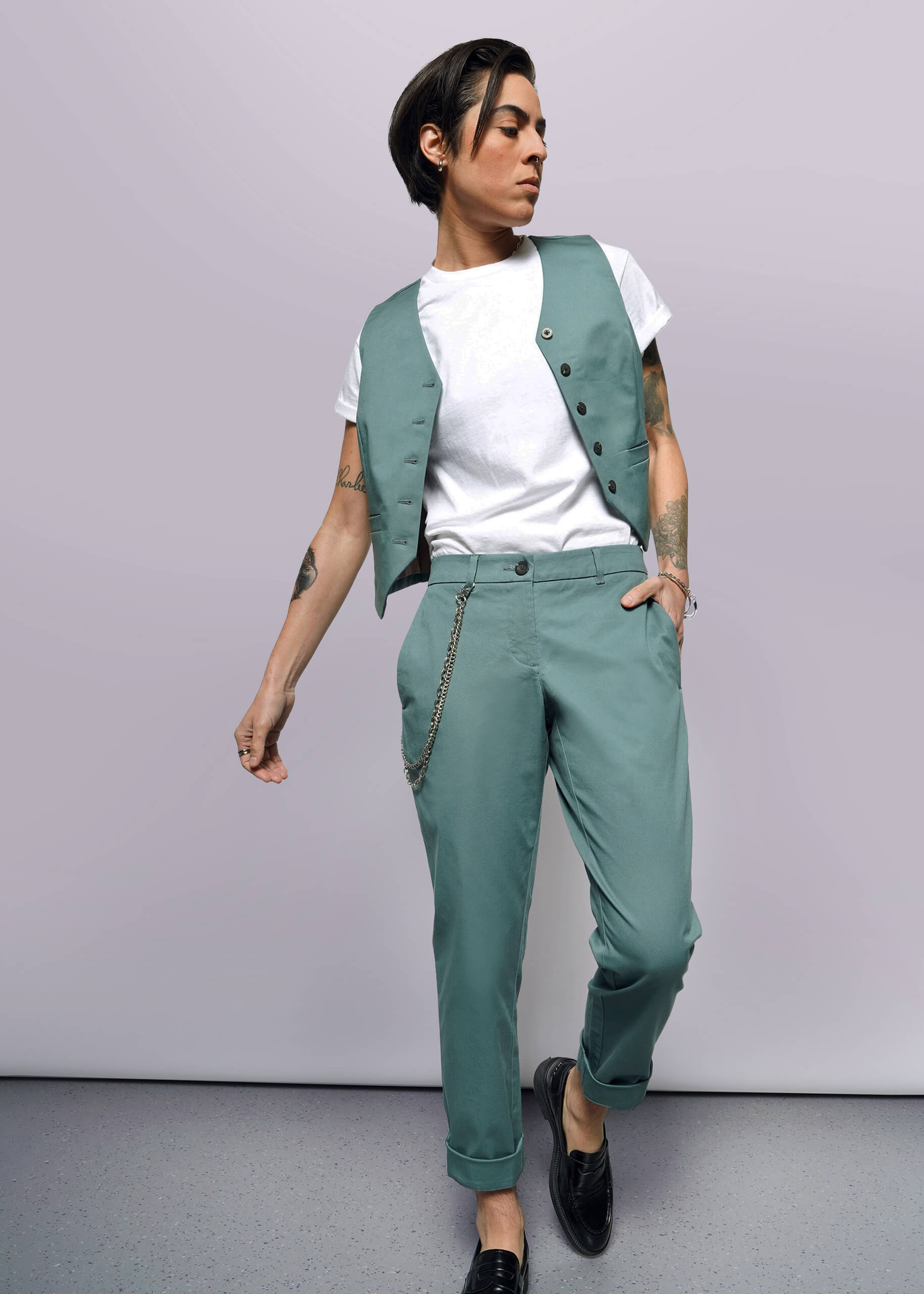 S=Person with short dark hair wears a white t-shirt, teal vest, and The Essential Trouser in a relaxed mid-rise fit with a chain accessory, standing against a plain white background and looking to the side.