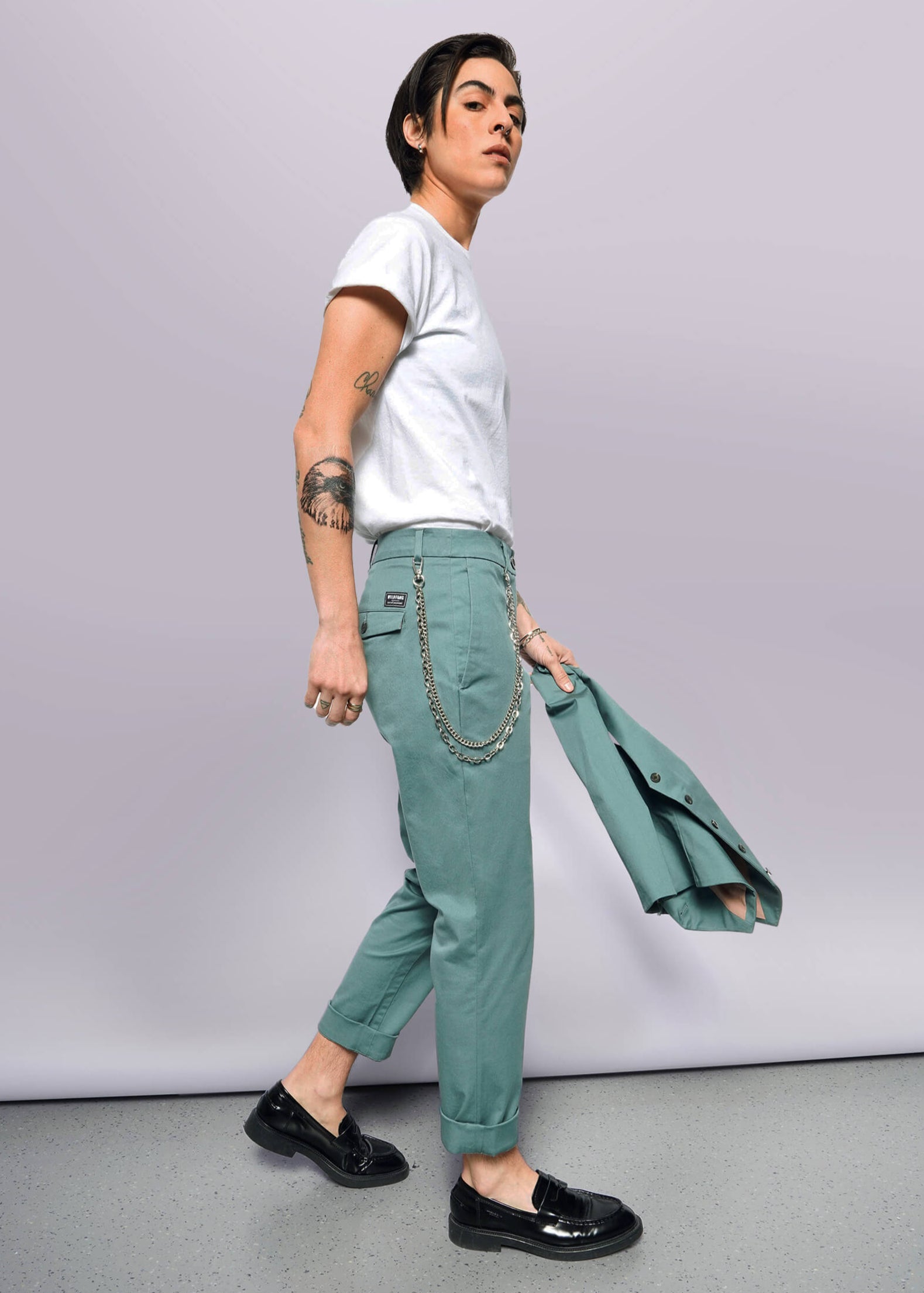 S=A tattooed person in a white t-shirt, The Essential Trouser in teal with a chain, black shoes, and holding a matching teal blazer walks against a plain white background.