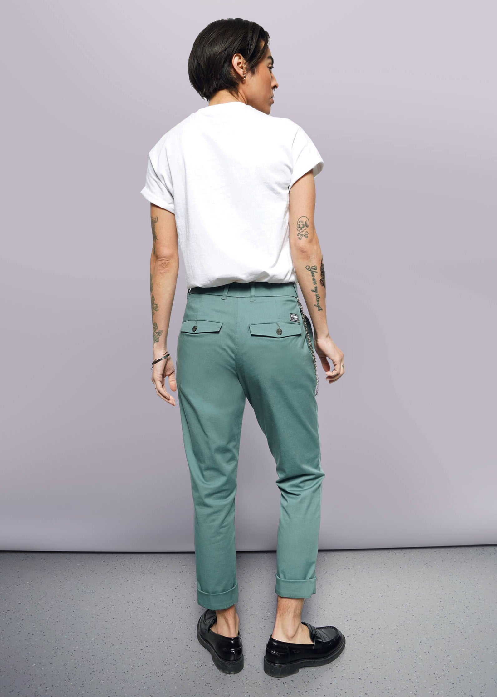 S= A person with short dark hair wearing a white rolled-sleeve tee, The Essential Trouser in teal with a relaxed mid-rise fit, and black loafers stands facing away. Tattoos are visible on both arms. The background is plain and well-lit.