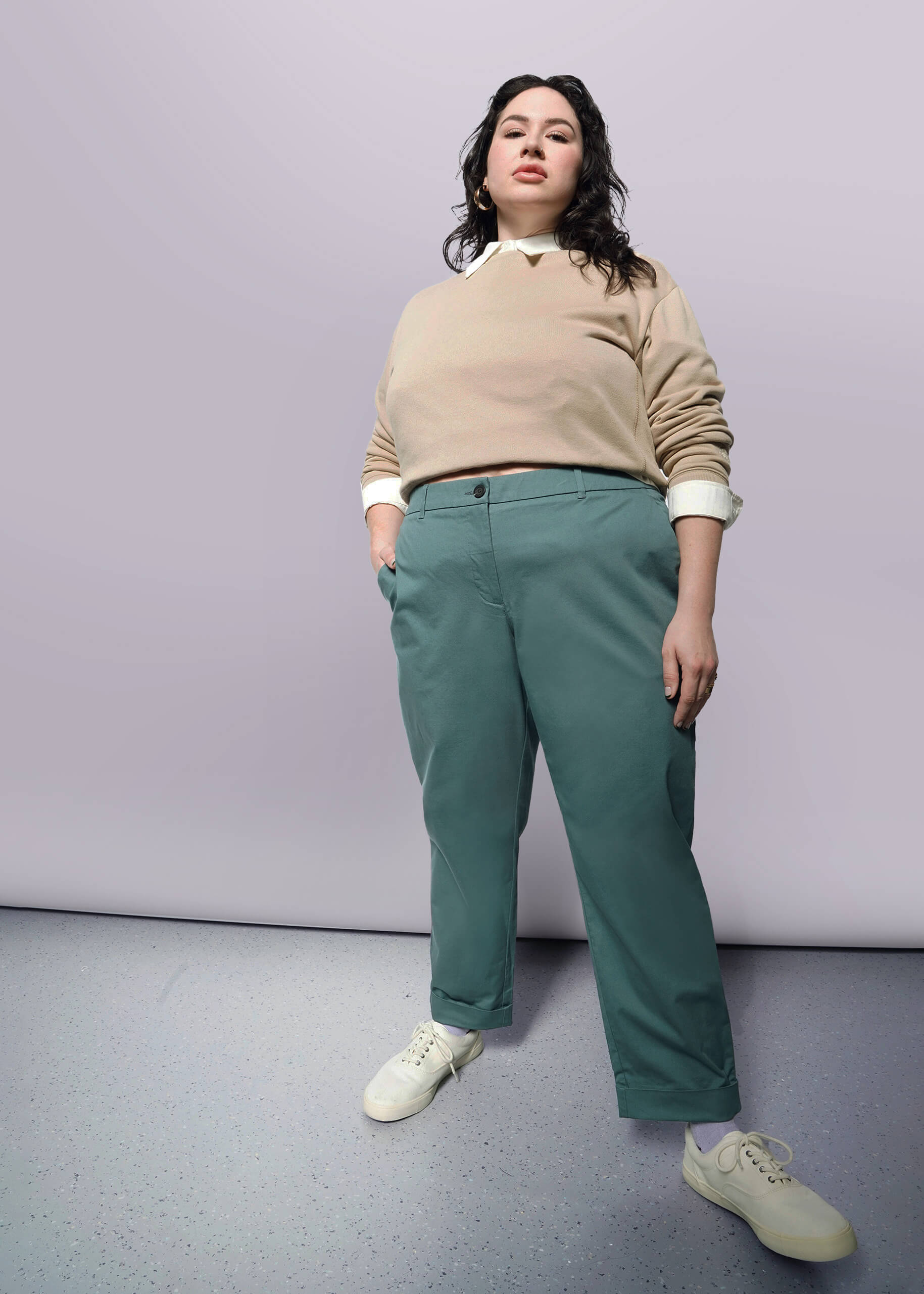 1X= A person with long dark hair stands confidently with one hand in their pocket, wearing a beige sweater, The Essential Trouser in teal with a relaxed mid rise fit, and white sneakers against a plain light background.