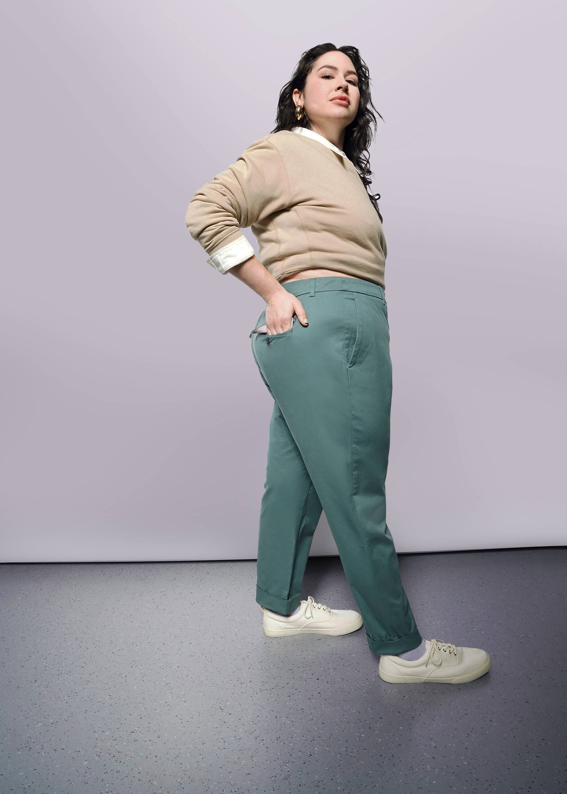 1X=A woman poses confidently in a studio against a white background, wearing The Essential Trouser in teal green with a relaxed mid-rise fit, paired with a beige sweater and white sneakers. She has dark wavy hair and one hand in her pocket.