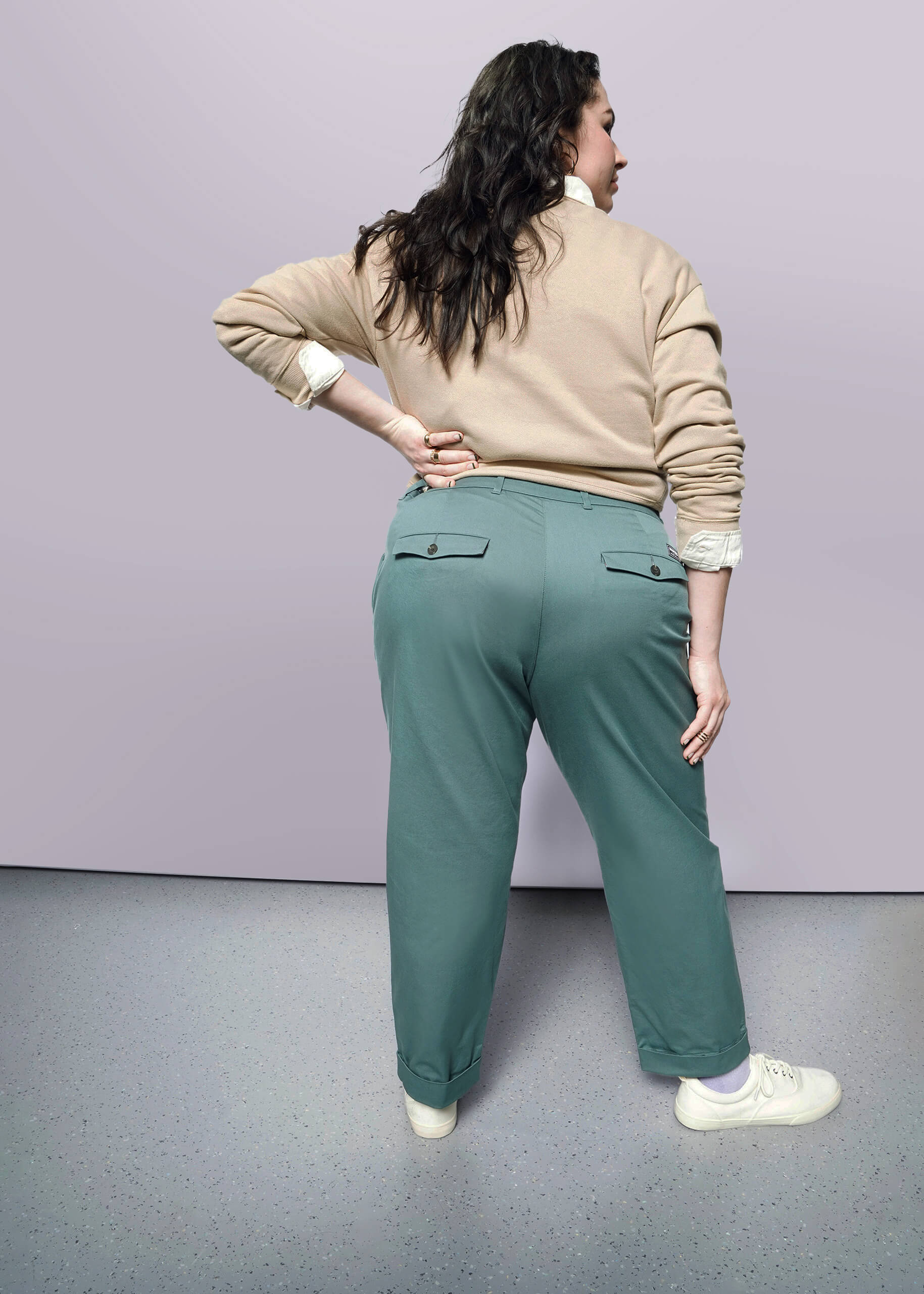 1X= A person with long dark hair, wearing a beige sweater and The Essential Trouser in a relaxed mid-rise fit, stands facing away with one hand on their hip against a light background.