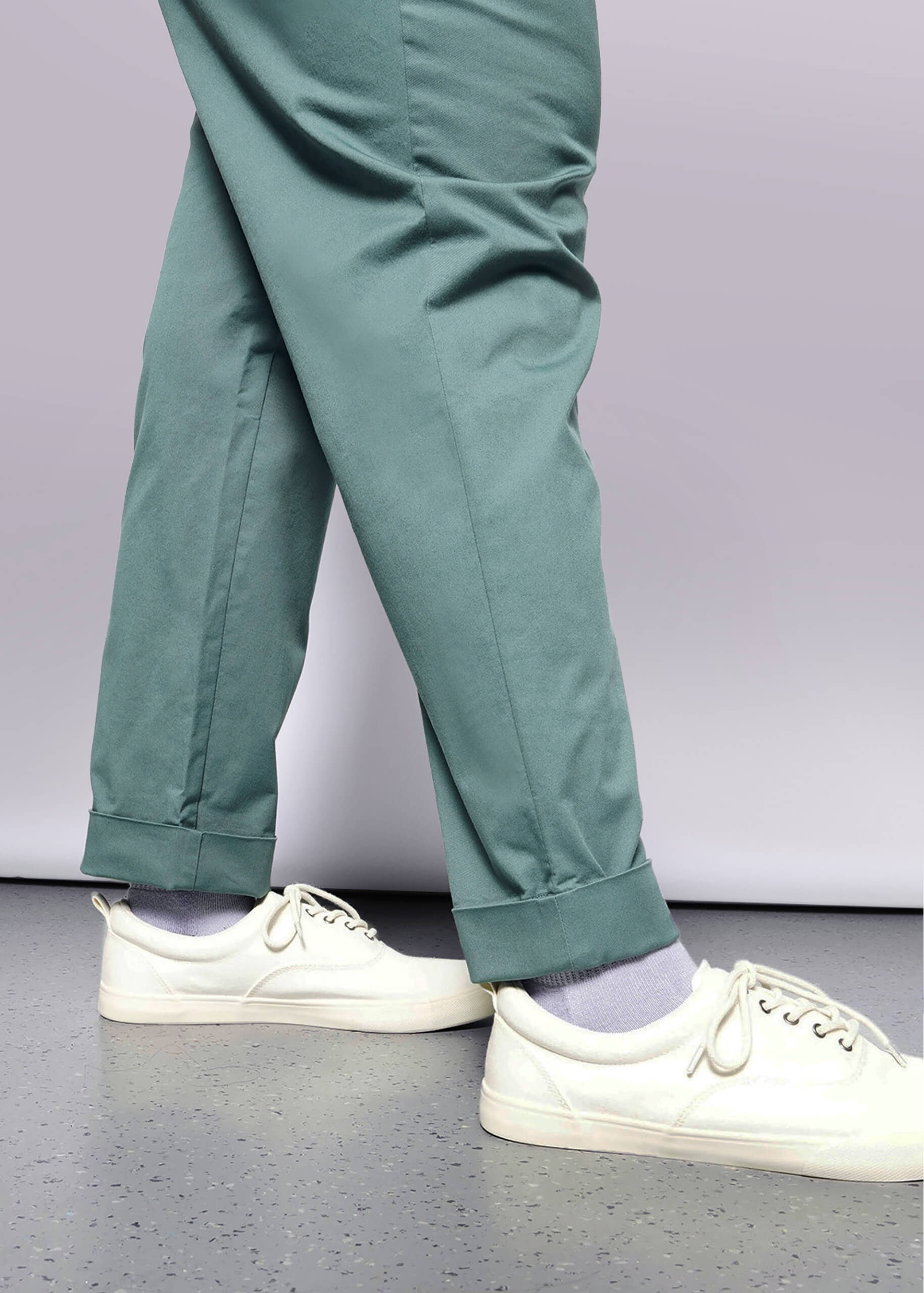 1X=A person wearing The Essential Trouser in teal with a relaxed mid-rise fit, paired with light purple socks and white sneakers, walks on a light floor. Only their legs and feet are visible against a white background.