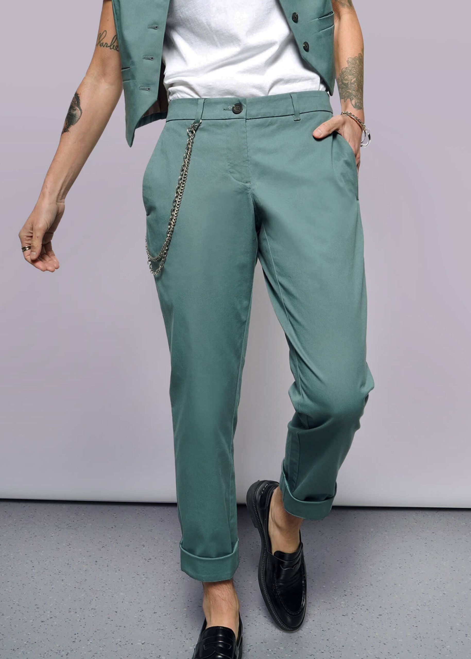 S= A person stands casually with a tattooed arm, hand in their pocket, wearing The Essential Trouser in classic teal with a chain accessory, paired with a white shirt and cropped teal vest. They wear black loafers against a plain white background.