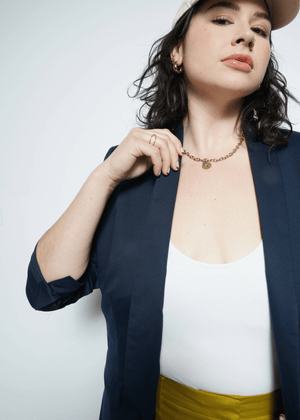 1X=A woman in The Essential Tux Blazer with rolled-up sleeves layers it over a white top and yellow pants. Shes styled with a beige cap, gold chain necklace, and hoop earrings. Her dark curly hair and confident gaze enhance the jackets tailored fit, perfectly complementing her style.
