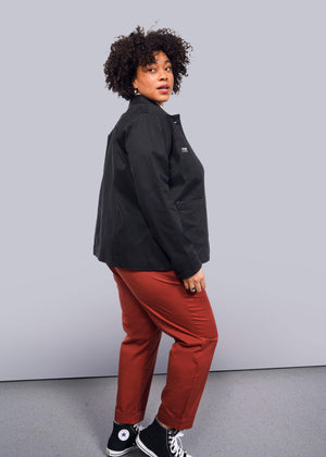 1X=A person with curly hair stands against a white background, wearing The Essential Utility Jacket and rust-colored pants. They glance over their shoulder and wear black sneakers.