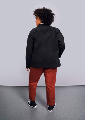 1X=A person with curly hair, dressed in the Essential Utility Jacket with deep pockets for convenience and stretch comfort for easy movement, red pants, and black sneakers with white soles, stands against a plain white background facing away from the camera.