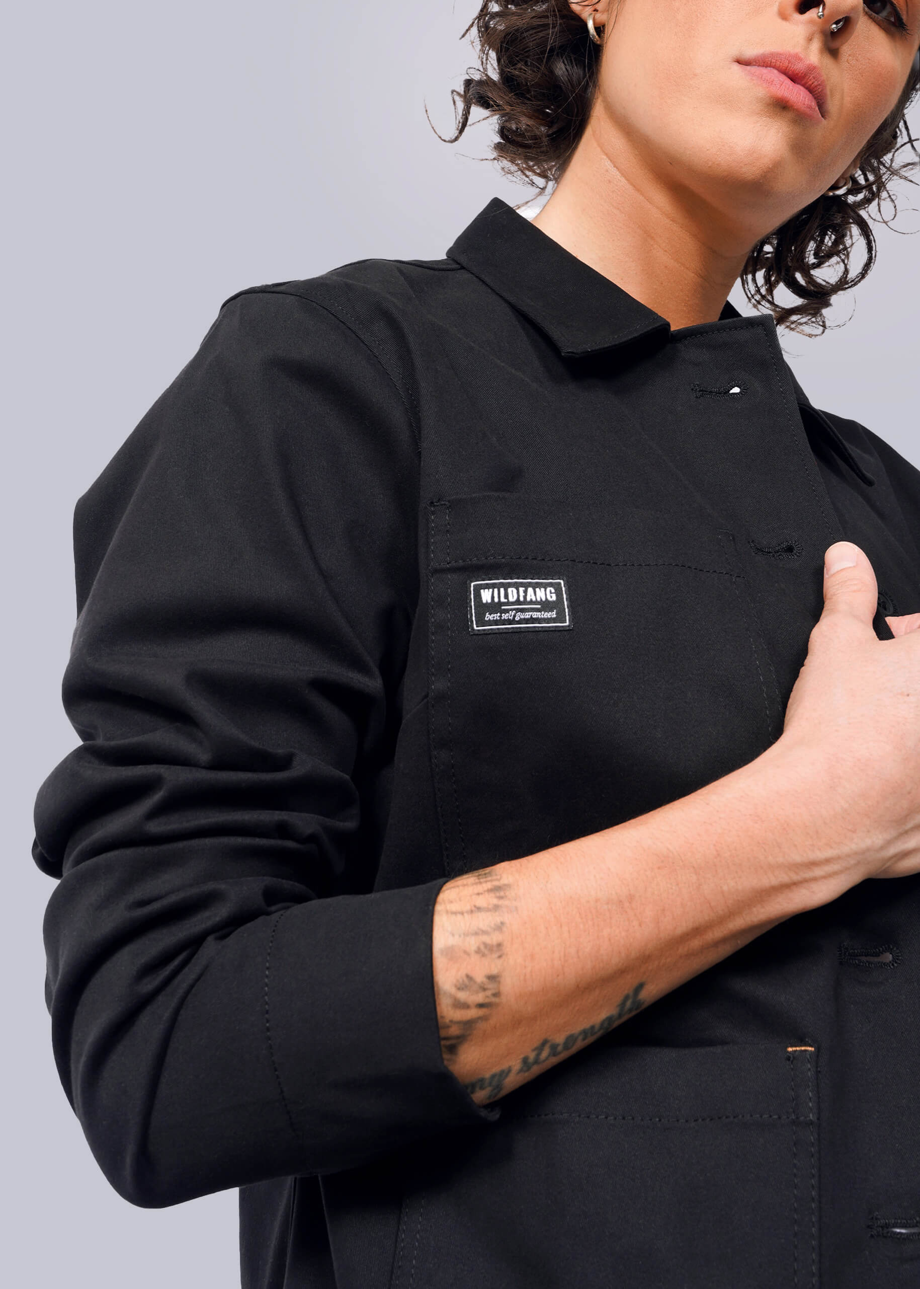 S=Against a plain background, a person models The Essential Utility Jacket with a Wildfang patch. The jacket features stretch comfort and deep pockets. With an arm partly raised, the intricate details of the jacket are highlighted alongside their forearm tattoo.