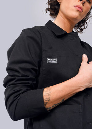 S=Against a plain background, a person models The Essential Utility Jacket with a Wildfang patch. The jacket features stretch comfort and deep pockets. With an arm partly raised, the intricate details of the jacket are highlighted alongside their forearm tattoo.