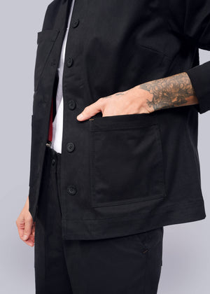S=A tattooed person wears The Essential Utility Jacket, slipping their hand into its large front pocket. This black jacket over a white shirt offers stretch comfort, combining style and practicality for those valuing deep pockets and fashion details.