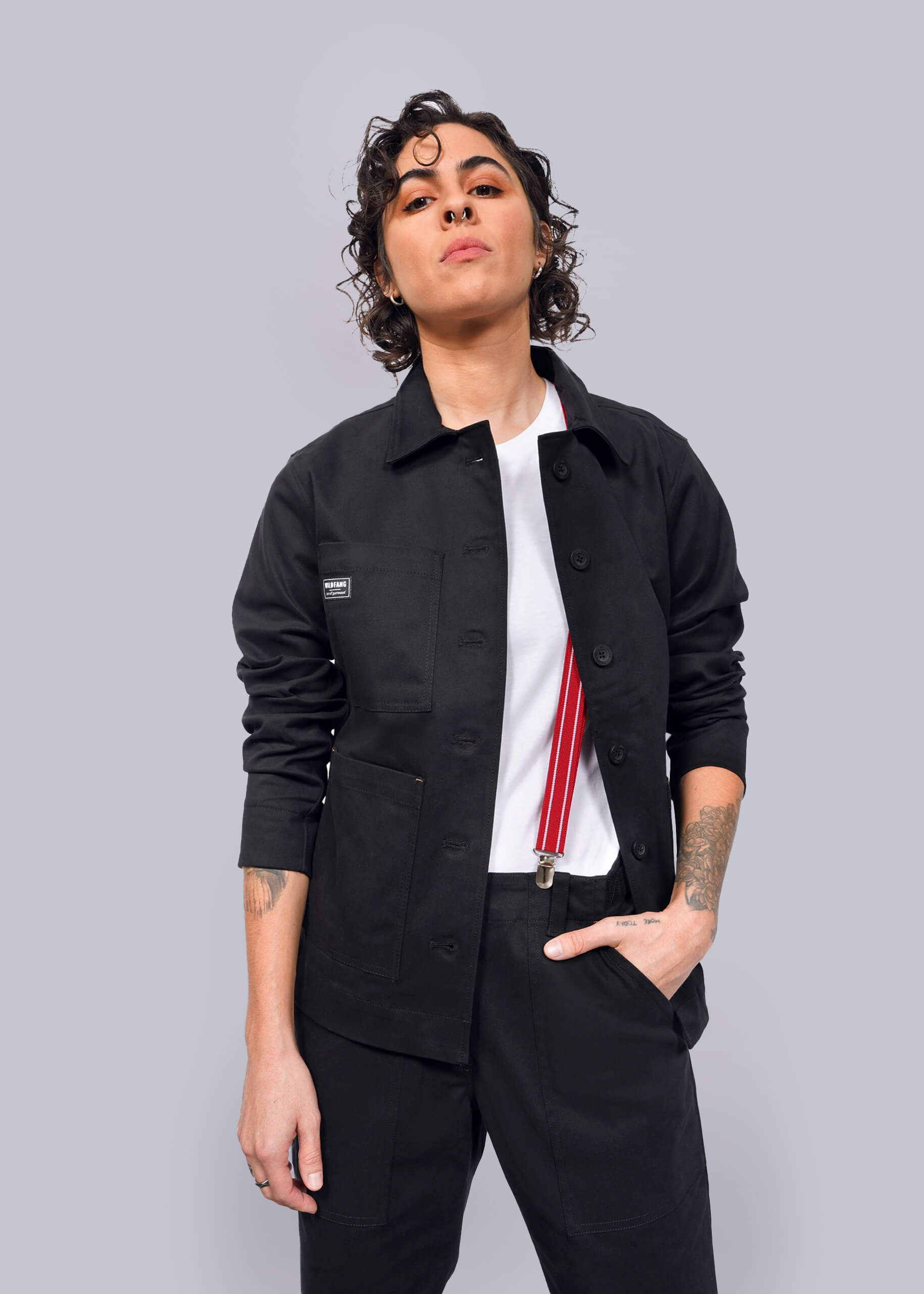 S=Wearing The Essential Utility Jacket and comfortable stretch pants, a person with short curly hair pairs their look with a white shirt and red suspenders, standing confidently against a white background.